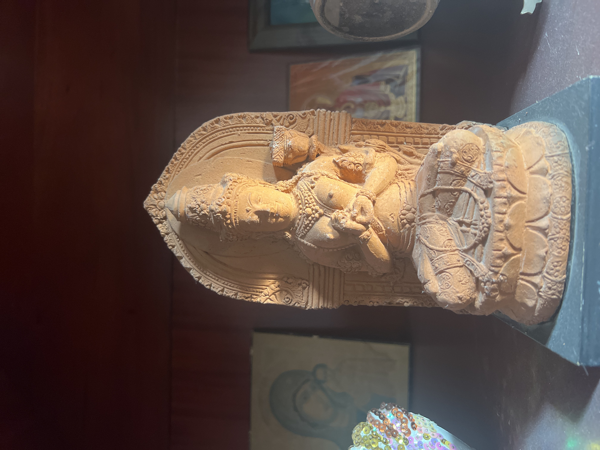 This is a photograph of a terracotta sculpture of a reclining figure, possibly a deity or royalty, set against a dark background. The sculpture depicts a person with elaborate headdress and jewelry, resting on a decorative base. The figure appears to be in a serene or contemplative pose. The sculpture is lit from above, creating highlights and shadows that emphasize the intricate details of the carving. In the background, there are hints of other artworks or objects, suggesting the sculpture is part of a collection or displayed in a home or gallery setting. The overall impression is one of cultural or artistic display, with a focus on the detailed craftsmanship of the terracotta piece. The image does not contain any visible text.