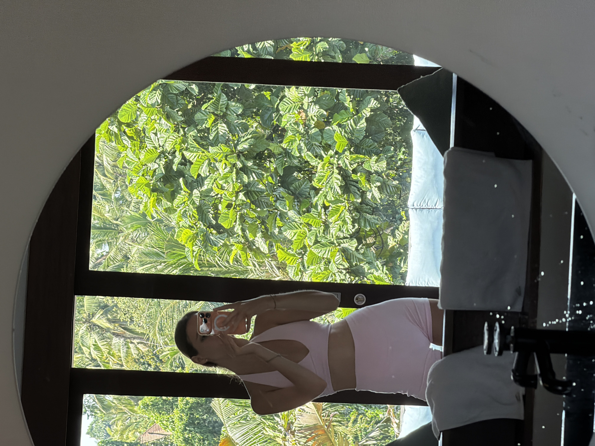 A woman is standing in what appears to be a bathroom and taking a selfie with her mobile phone. She is wearing a light pink two-piece athletic outfit. The background includes a window with a view of lush tropical vegetation. Based on the information provided, the location of the image is Petulu, Indonesia.
