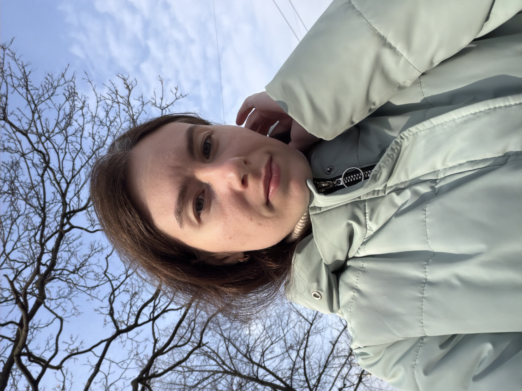 A selfie captures a woman wearing a light green quilted jacket outdoors, with a tree branch silhouette against a blue sky and some cloud cover in the backdrop. She is holding her hand near her ear and looks directly at the camera.
The location appears to be a park or outdoor area, possibly within the city of Saint Petersburg, Russia, based on the tree species and general ambiance.