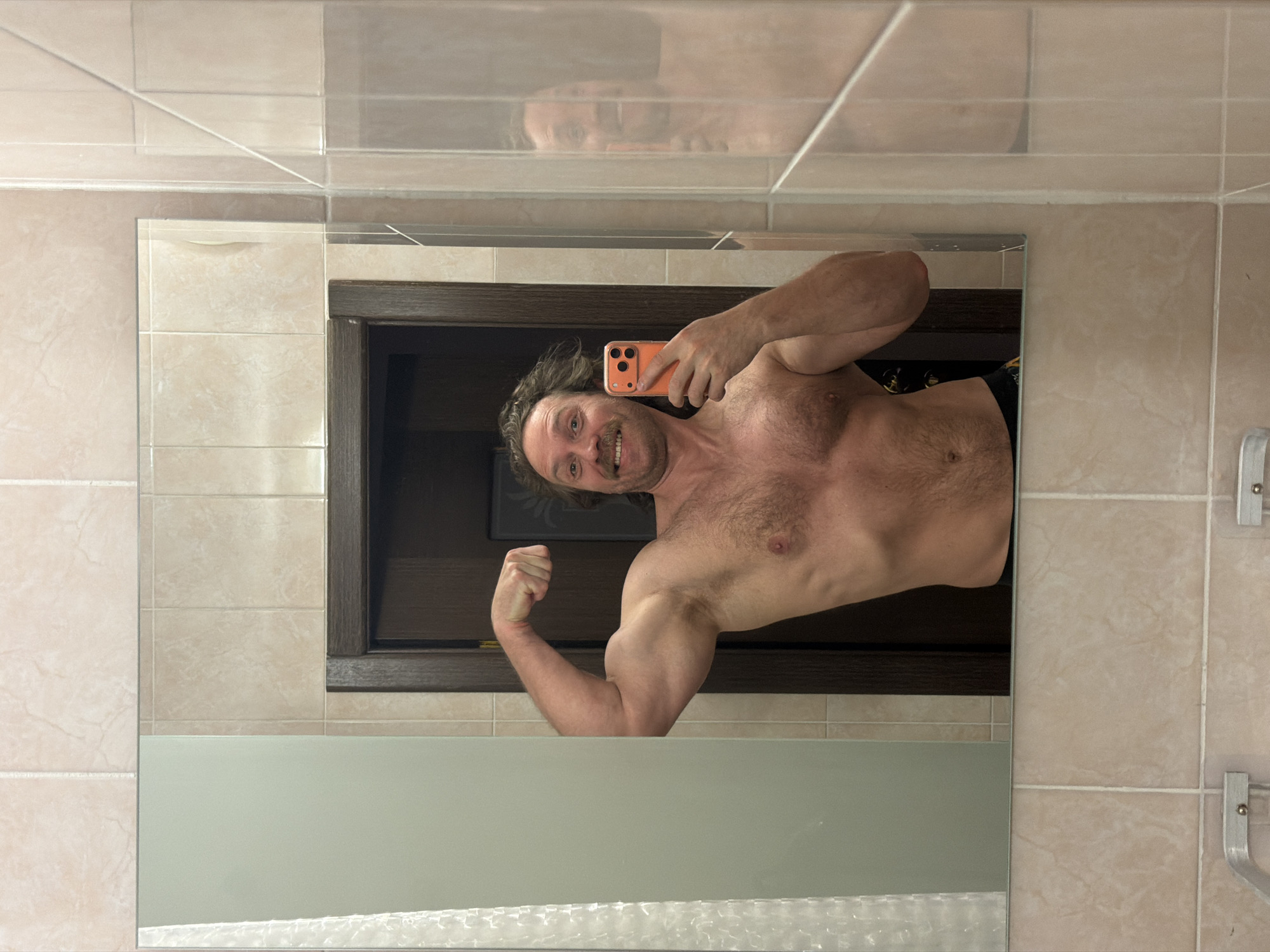 A man is reflected in a mirror in a bathroom. The man, fair-skinned with dark hair and a mustache, is holding a phone in his right hand, presumably taking a selfie. He is shirtless and flexing his left bicep. The bathroom features light beige tiles on the walls, a dark brown frame around the mirror, and what appears to be a bathtub below the visible portion of the mirror. The location is not discernible from the image.