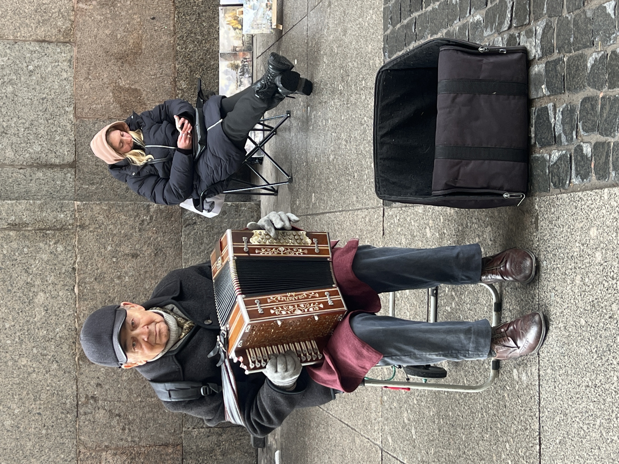 The image shows a man playing an accordion on a sidewalk in what appears to be St. Petersburg, Russia, based on the architectural style of the buildings depicted in the paintings in the background. The man is seated on a small folding chair, wearing a dark coat, grey trousers, and brown leather shoes. He is holding the accordion with both hands, which are covered in grey gloves, and is facing the camera. A black bag is next to him. Behind him, a young woman in a hooded jacket is seated on another folding chair. She is likely a companion or relative of the man. The setting is a city sidewalk, with cobblestone on one side and stone slabs on the other. There are several paintings on display near the woman. The overall impression is that of a street musician performing in a public space.