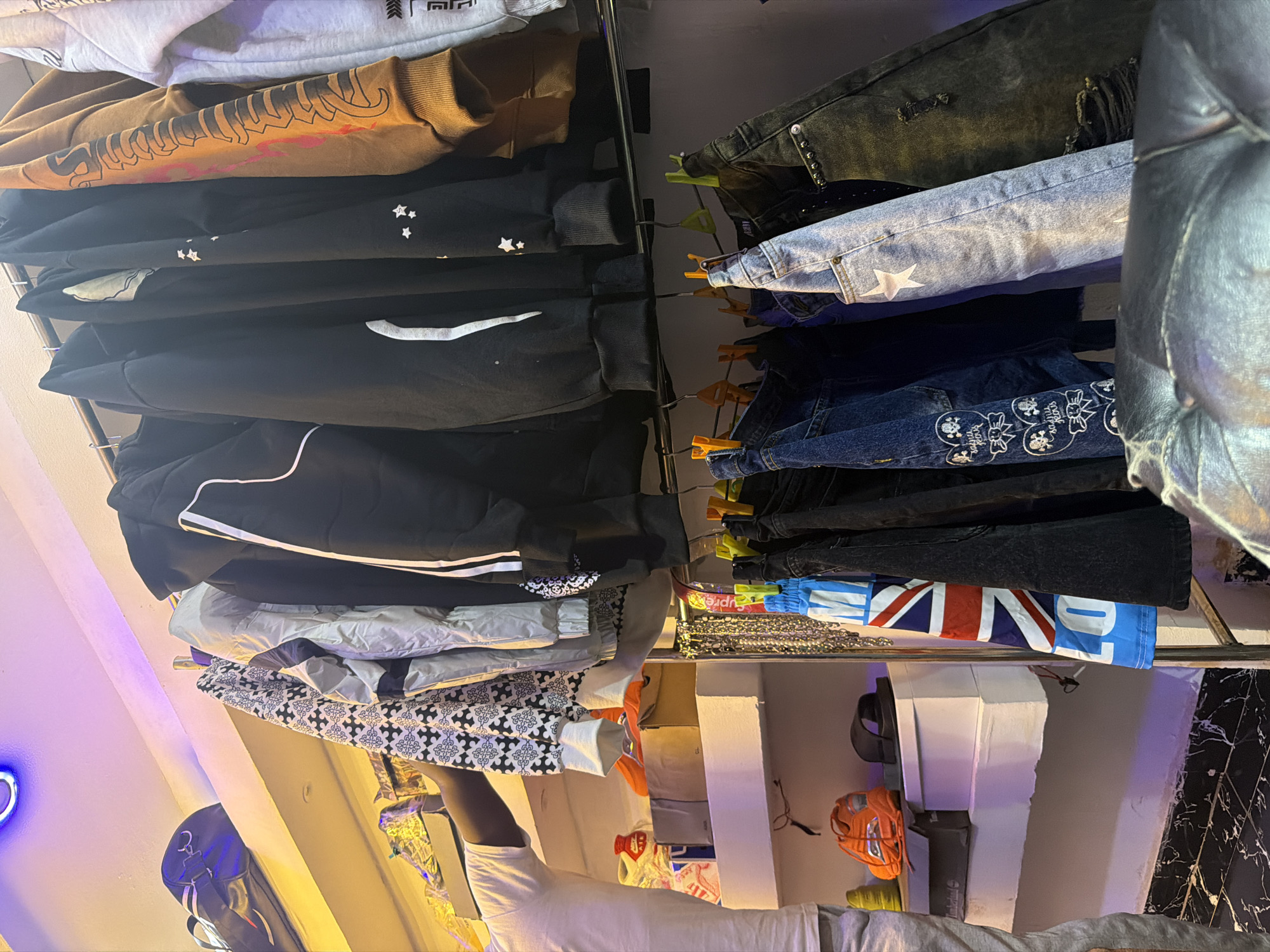 The image depicts an indoor retail setting, likely a clothing store, in Bukuru, Nigeria. A person, partially visible from the left, is reaching towards a rack of clothing. The rack holds several garments, including hoodies and what appear to be jeans. Some of the clothing items feature distinctive patterns and designs, such as a black hoodie with white stripes, a patterned jacket, and jeans with the Union Jack design. On shelves below the clothing rack, various items are displayed, including shoes, boxes, and what seem to be accessories like orange slides. The environment is illuminated by artificial lighting, with purple and yellow hues visible on the walls and ceiling. The overall scene suggests a commercial space focused on apparel sales.