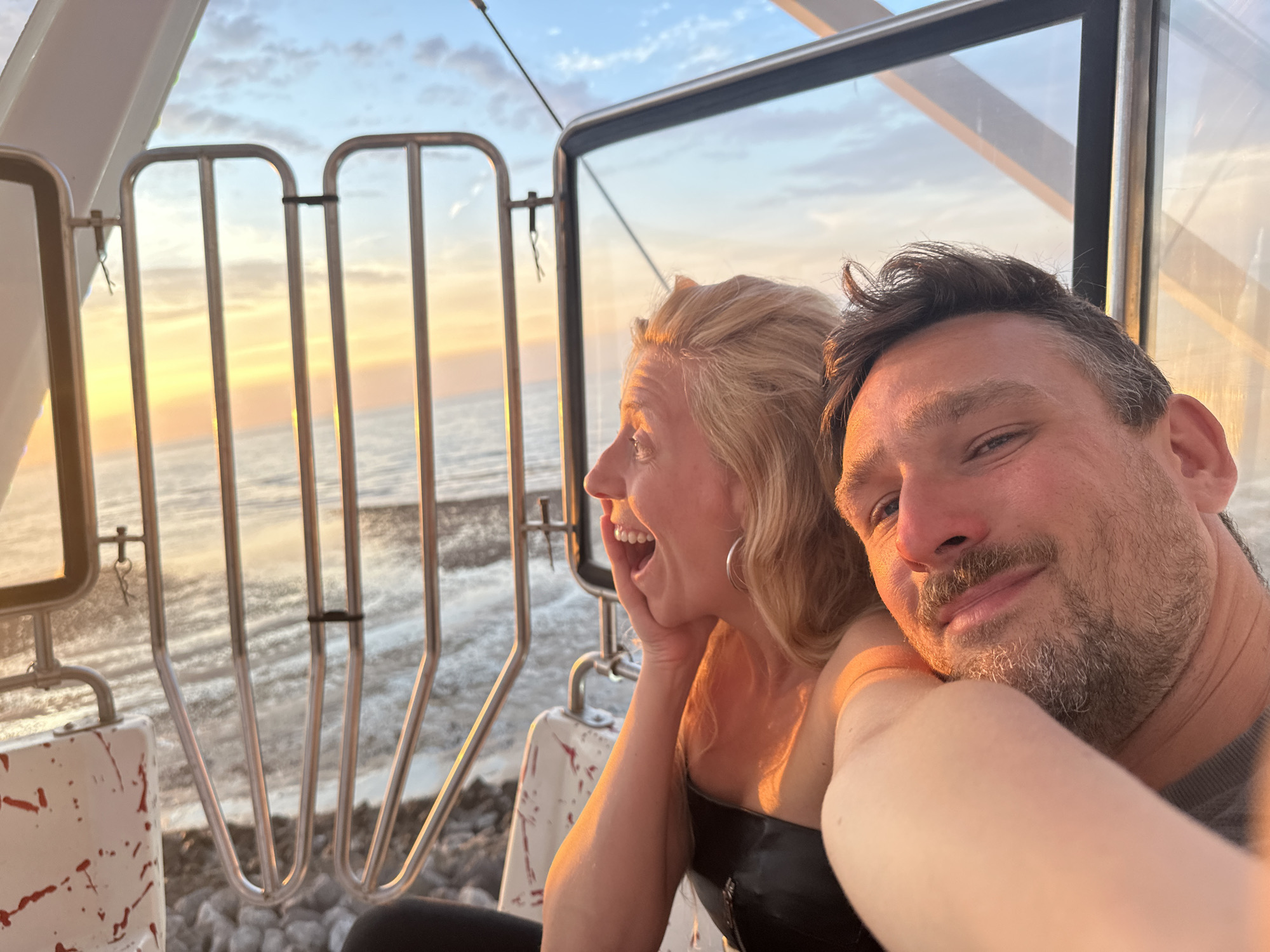 In Le Tréport, France, a couple is captured in a selfie taken from within a gondola, likely part of the Funiculaire du Tréport. The setting sun casts a warm glow over the scene, highlighting the couple's faces and the surrounding environment. The woman, positioned to the left, has a surprised expression, her hand resting on her chin, and her mouth open in a wide smile. She is wearing a black strapless top. The man, on the right, is looking at the camera with a relaxed smile. He has a beard and is wearing a grey shirt. The gondola's metal railing and a portion of its white interior with red splatters are visible. Through the glass window of the gondola, the sea and the sky are visible, with the sunset creating streaks of orange, yellow, and blue. The water appears choppy, and a rocky shore is visible in the foreground.