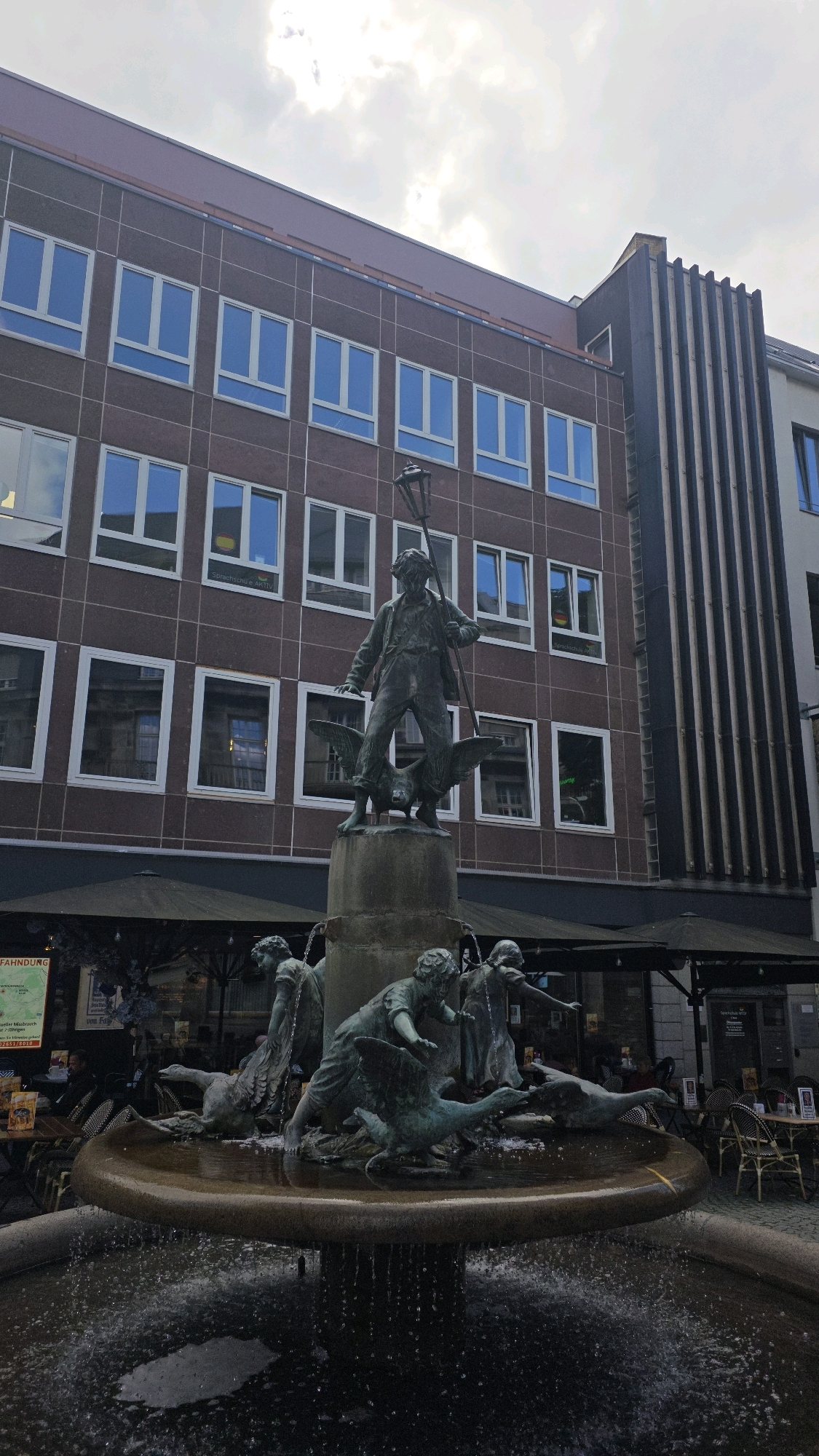 The image depicts the "Rheinlandlied" fountain located in Bonn, Germany. The fountain features a central bronze sculpture of a man holding a lantern pole, standing on a base with two winged creatures, possibly geese or swans. Around the base of the fountain, there are other bronze figures, including children and more winged creatures, interacting with the water. The fountain is set in a courtyard with outdoor seating areas for cafes or restaurants, indicated by tables and chairs. In the background, there is a multi-story building with a patterned facade and numerous windows. The sky is overcast.