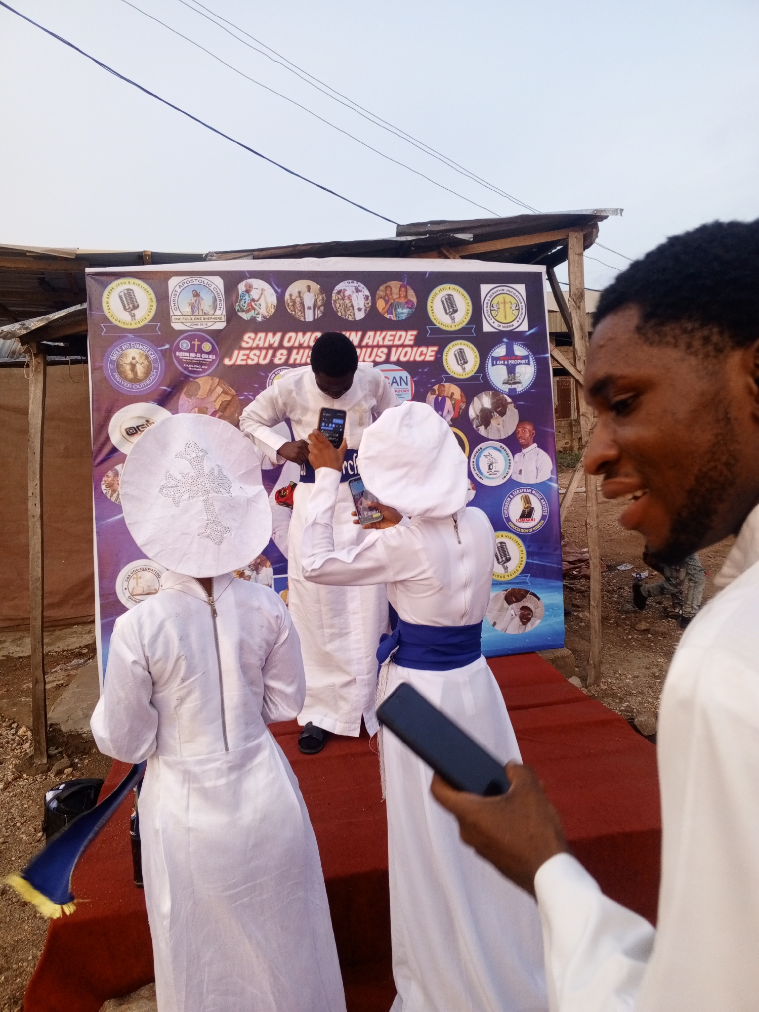 Here is a detailed description of the image:
**Scene:** The photograph captures an outdoor event or gathering. The setting appears to be a public space in a city. The background includes an informal structure, possibly a temporary or makeshift building, indicating the location may be a less developed area.
**People:** The photo features several individuals. Two people are wearing white robes, a white hat, and a blue sash, possibly religious garments, with one person facing away from the camera with a cross decoration on their hat. Another person dressed similarly stands partially visible behind and between the other two. A fourth person, wearing a white robe, stands to the right and is holding a phone and appears to be smiling. Another person is positioned in the background taking a photo with their phone.
**Objects:** A large banner stands behind the group, displaying text and several circular images, potentially logos or portraits. The text on the banner includes "SAM OMON AKEDE JESU & HIS VOICE." In the foreground, one of the people holds a mobile phone.
**Actions & Interactions:** The people are gathered around the banner. One person appears to be taking a photograph or recording a video with a mobile phone, while the others may be posing or presenting for the camera. The person on the right appears to be looking at the phone and smiling.
**Environment:** The surroundings include unpaved ground with sparse vegetation and gravel. The lighting suggests it is either early morning or late afternoon.