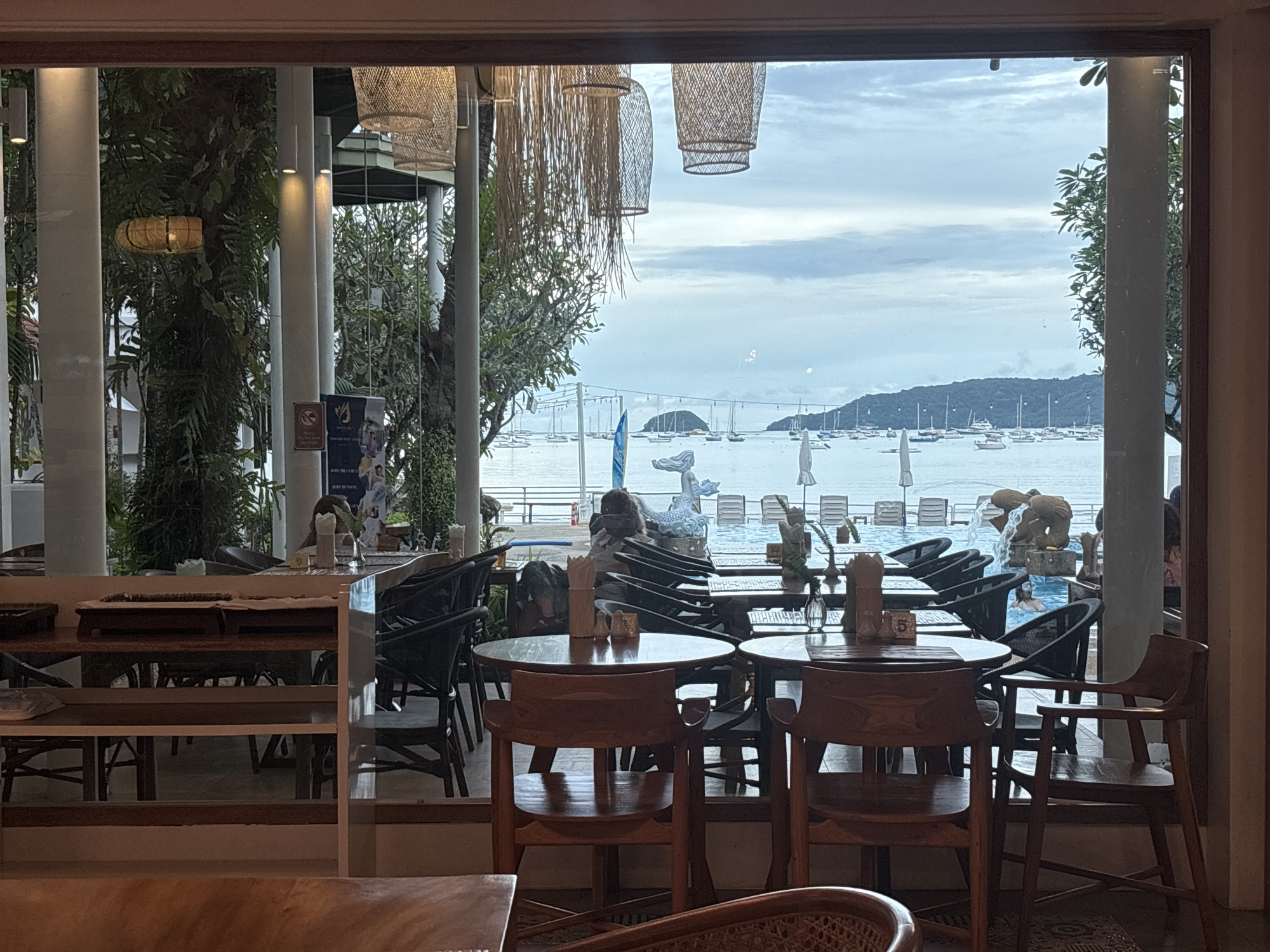 The image shows an indoor restaurant with a view of a bay, likely in Chalong, Thailand. The restaurant has large windows and is furnished with wooden tables and chairs. Hanging lamps made of woven material are visible. Outside, there is a body of water with numerous boats docked. The sky is overcast, and a distant island is visible. In the foreground, several tables are set up, some with salt and pepper shakers. A few people are seated at tables, and some are seen walking around. In the background, through the windows, a swimming pool area with lounge chairs is visible, and one person is in the pool. The overall scene depicts a relaxed atmosphere in a seaside establishment.