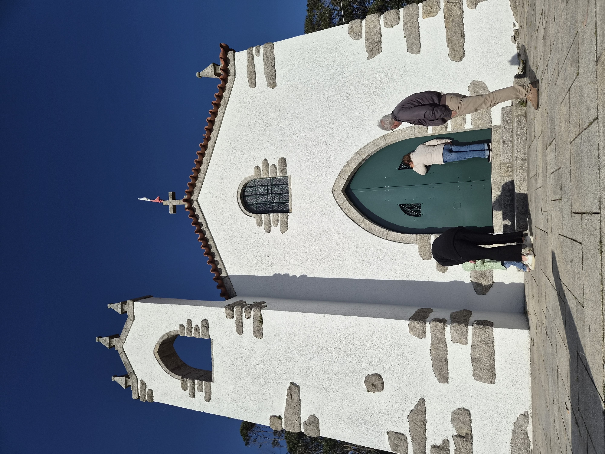 The image depicts the Igreja de São João Baptista, a church in Esposende, Portugal. The church is a white building with stone trim, a red-tiled roof, and a cross on top. Several people are standing in front of the church's green arched doors. One person, wearing khaki pants and a dark jacket, appears to be looking at the doors. Another person, wearing blue jeans and a white jacket, is also looking at the doors. A third person, wearing black pants and a green shirt, is also present. The sky is clear and blue. The church is located on a stone surface.