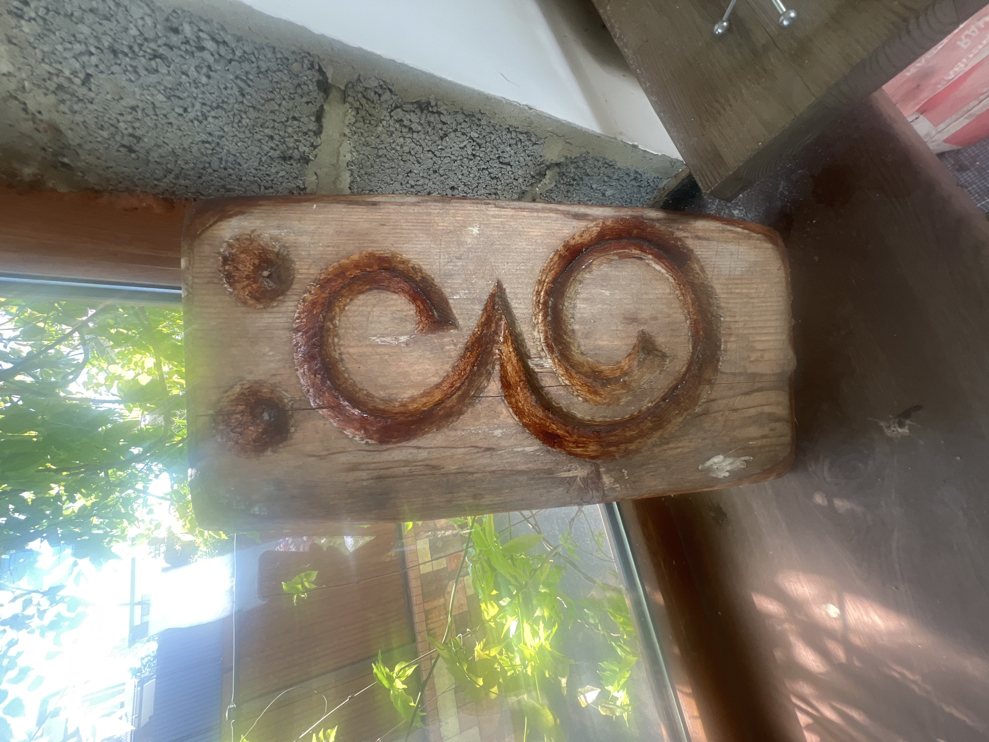 A wooden sign is mounted on a window frame in an outdoor setting. The sign is rectangular and features a carving of two interconnected, stylized spirals. Two circular, darker elements are located on the left side of the sign. The sign's location is likely in Sochi, Russia. A window with sunlight and green vegetation is visible below the sign. A wall is seen on the top.