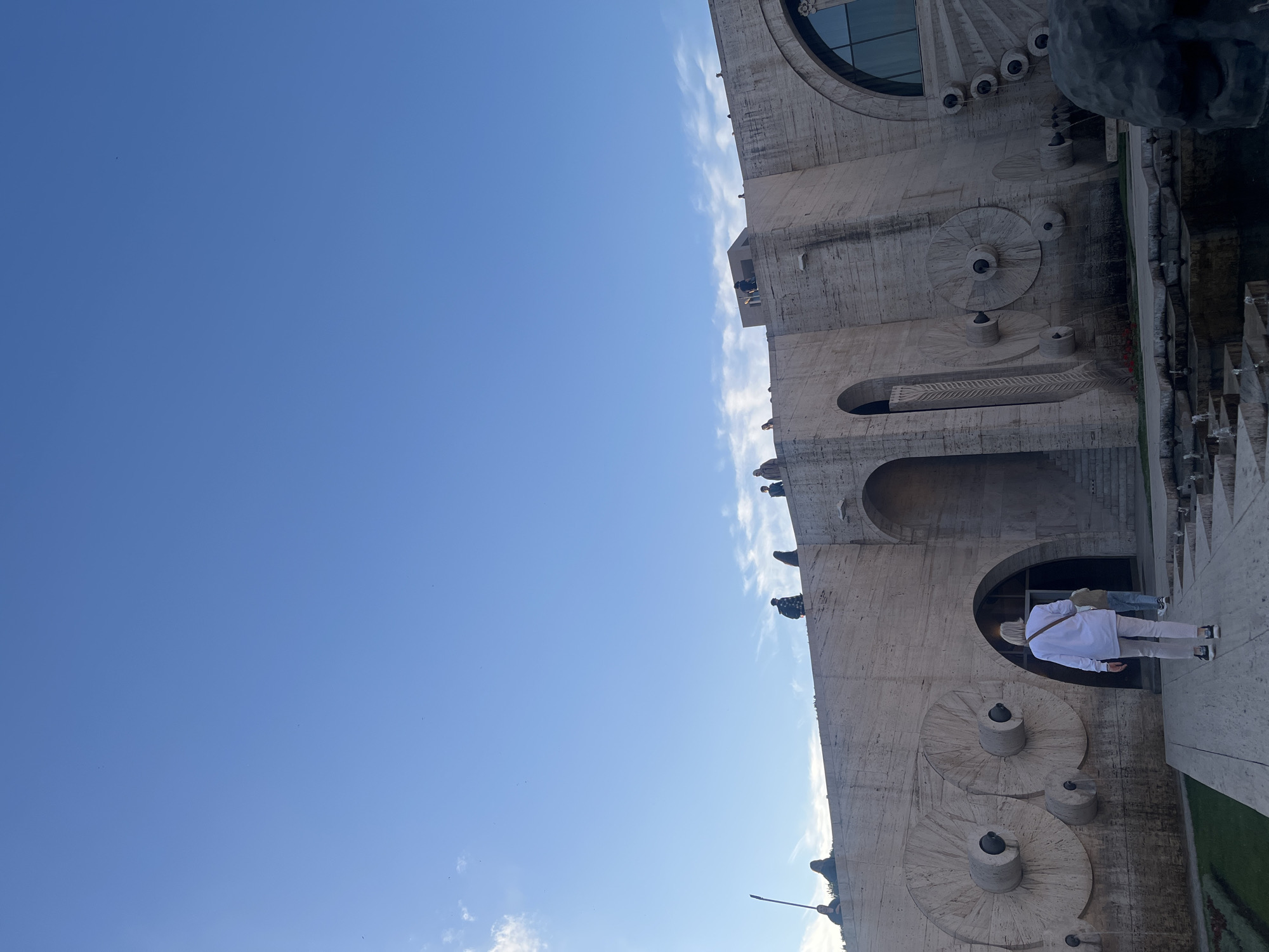 The image shows a partial view of the Cascade in Yerevan, Armenia, from a low-angle perspective. The Cascade is a massive stairway adorned with sculptures and fountains, built into a hillside. The structure is made of light-colored stone. In the foreground, a person in a white shirt and light pants is entering one of the arched openings. Above, there are various levels with sculptures and decorative elements. The sky is mostly clear blue, with a few wispy clouds.