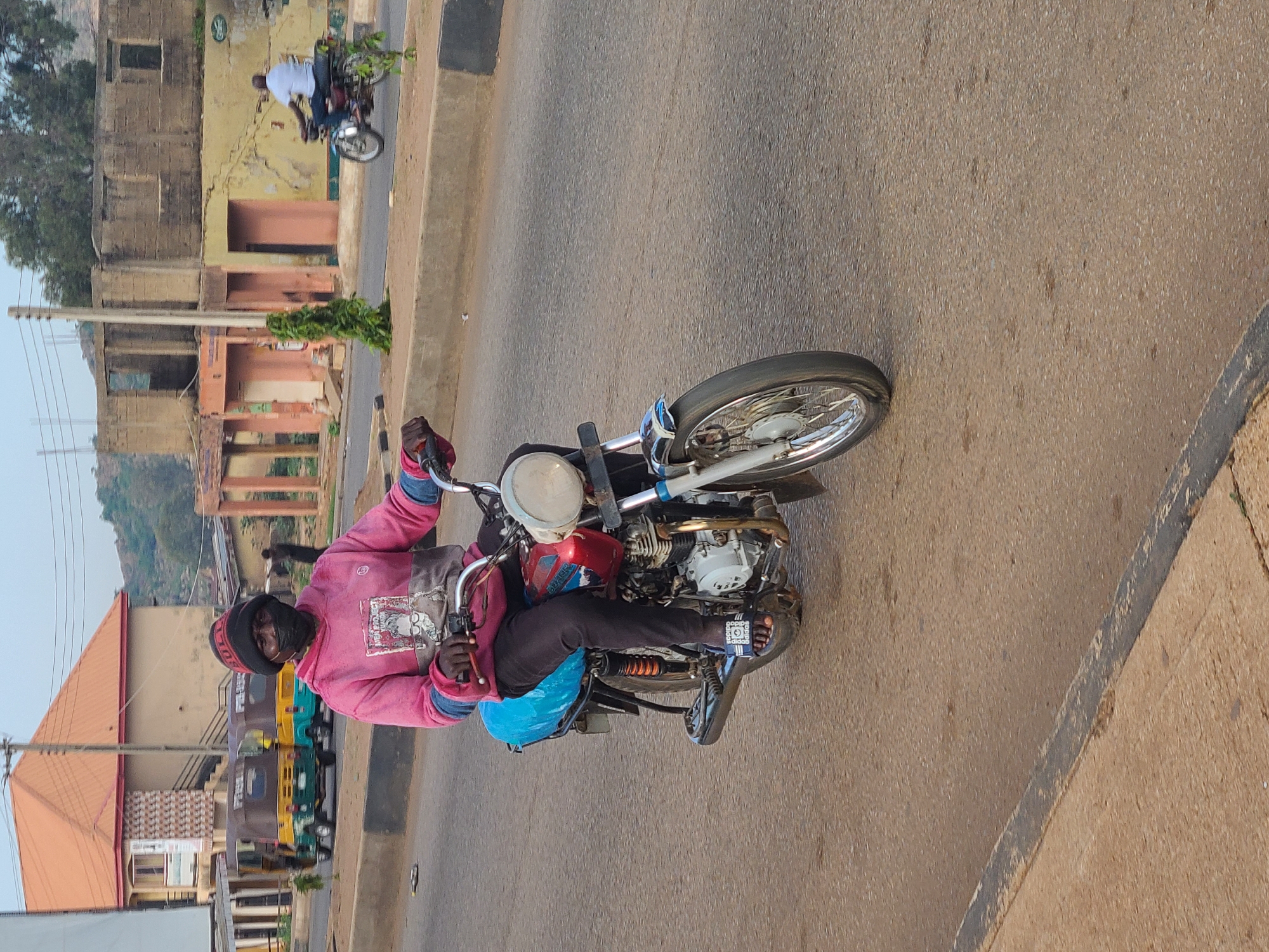A man rides a motorcycle on a paved road in Gura Top, Nigeria. He wears a pink sweatshirt, black pants, and sandals. A black hat and face covering obscure his face. The motorcycle is older and has a red and silver tank. A blue bag is strapped behind the seat. In the background are buildings, some dilapidated, and trees. Other motorcycles or vehicles are visible further down the road. The day appears bright.