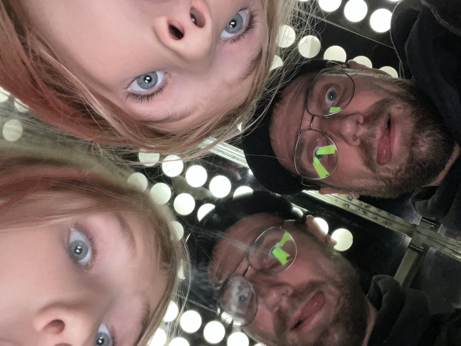 Two individuals, one adult male and one child, are seen in a mirrored surface. The image is taken from a low angle, looking upwards at their faces. The child is fair-skinned with blue eyes and light blonde hair. The male has a beard, glasses, and a dark cap. A green object is affixed to the glasses. Behind them, there are white circles illuminated. The mirrored effect creates a duplicate image of both individuals and the background.