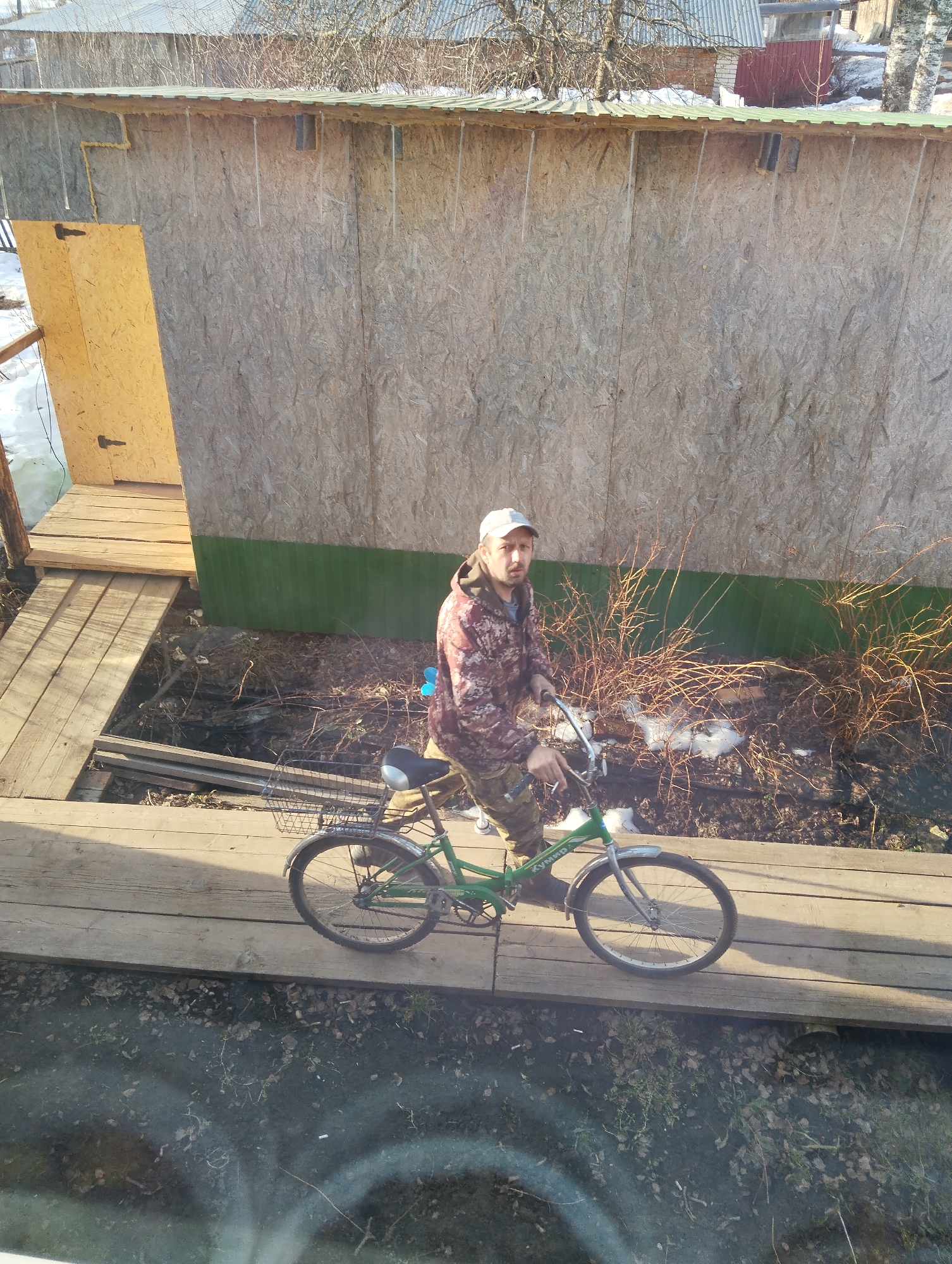 The image, likely taken from a window, shows a man on a green bicycle standing on a wooden walkway or bridge. He is wearing camouflage clothing and a baseball cap. Behind him is a wooden structure with a green painted base. Bare bushes are visible between the walkway and the structure. The ground is a mix of dirt, leaves, and some snow. The location is indicated as Krasnogorskoye, Russia.