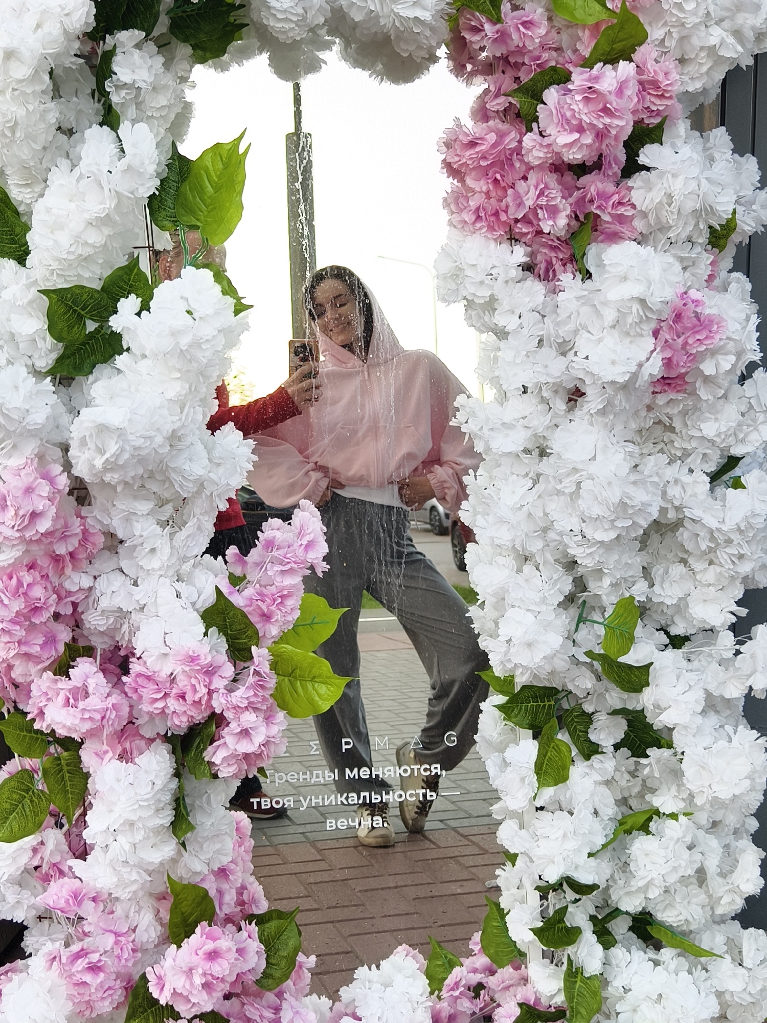 A woman in a pink hoodie and gray pants stands in front of a mirror decorated with white and pink flowers in what appears to be Tyumen, Russia. The mirror reflects her holding a cell phone, presumably taking a picture, while water streams down its surface. Part of the reflection also captures a person in a red shirt holding the mirror. Below the woman in the reflection, text in Russian is visible on the mirror's surface: "Тренды меняются, твоя уникальность вечна" (Trends change, your uniqueness is eternal). The ground is brick.