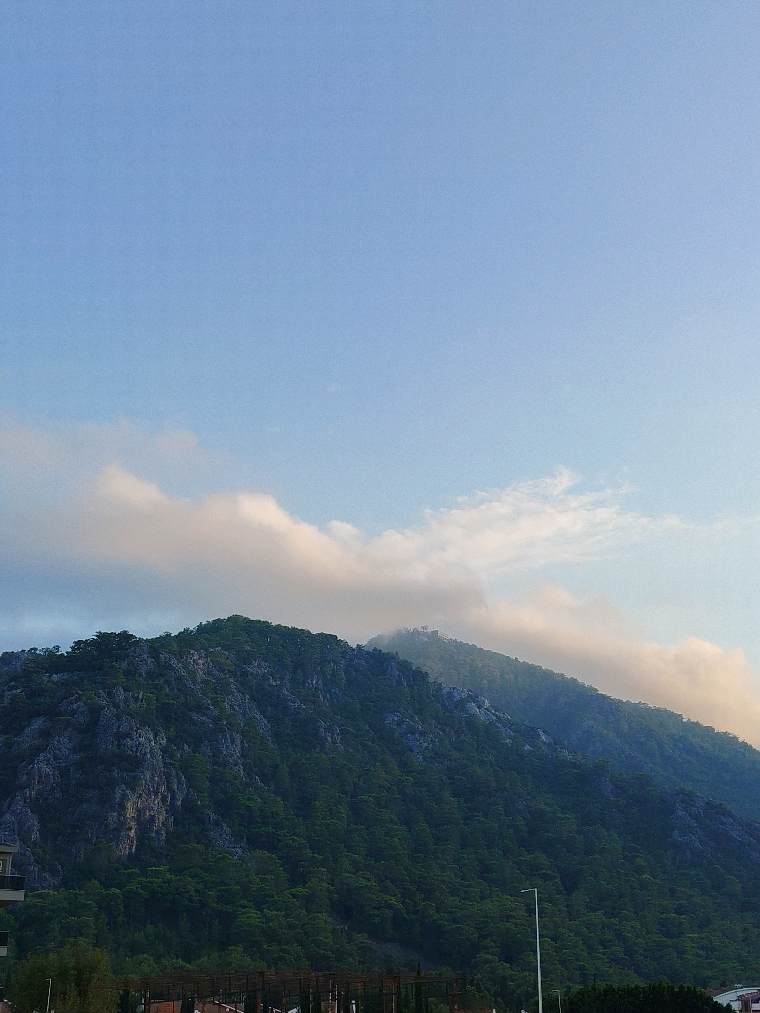 The image shows a panoramic view of a lush green mountainside in Konyaaltı, Turkey, with patches of rocky terrain visible. The mountain is densely covered in evergreen trees. A few wispy clouds are present in the sky, with some tinged with a warm, golden hue, suggesting either sunrise or sunset. The sky is a clear, pale blue. At the lower edge of the image, there are glimpses of buildings and structures, including a street lamp and some fencing, indicating a proximity to a developed area. The overall scene conveys a natural landscape juxtaposed with human habitation.