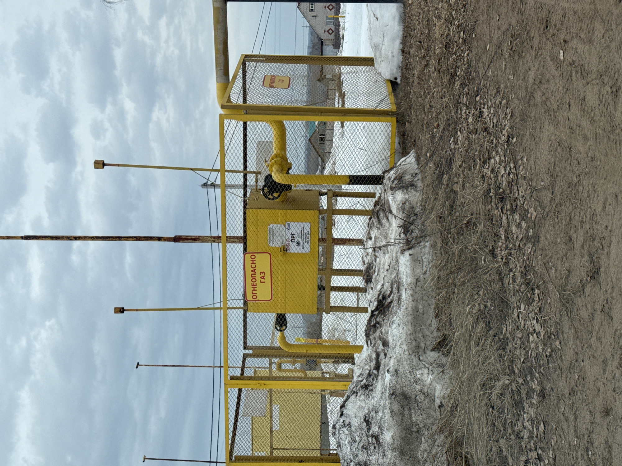 The image shows a yellow gas regulation point secured by a metal fence in Noviy, Russia. A sign on the gas regulation point reads "ОГНЕОПАСНО ГАЗ" which translates to "FLAMMABLE GAS" in English. There are a few houses in the background, and a patch of dirty snow is visible near the fence. The weather appears overcast. The surrounding ground consists of dirt, withered leaves, and dry grass.