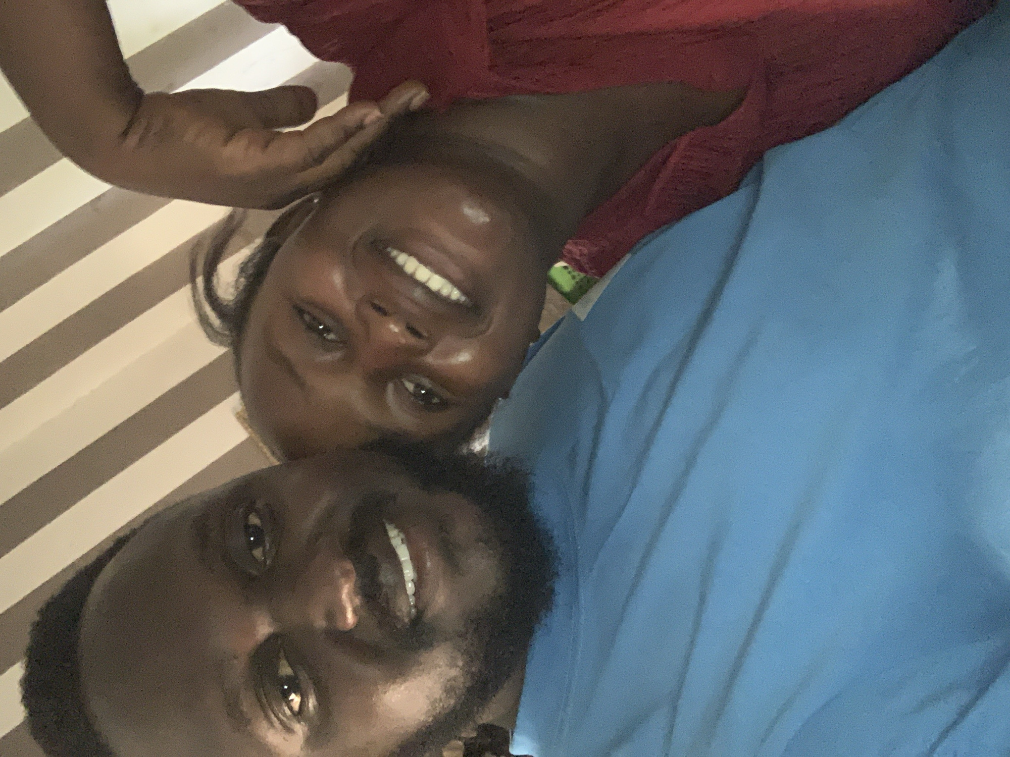 A close-up selfie shows two individuals, a man and a woman, in what appears to be an indoor setting. The man is on the left, wearing a blue t-shirt and has a beard and dark hair. He is smiling. The woman is on the right, wearing a red top. She is touching her own face with her right hand and smiling. Her hair is dark. The background features a striped pattern of beige and brown. The overall lighting is warm and soft. The image does not contain any discernible elements that would indicate the location as Jos, Nigeria.