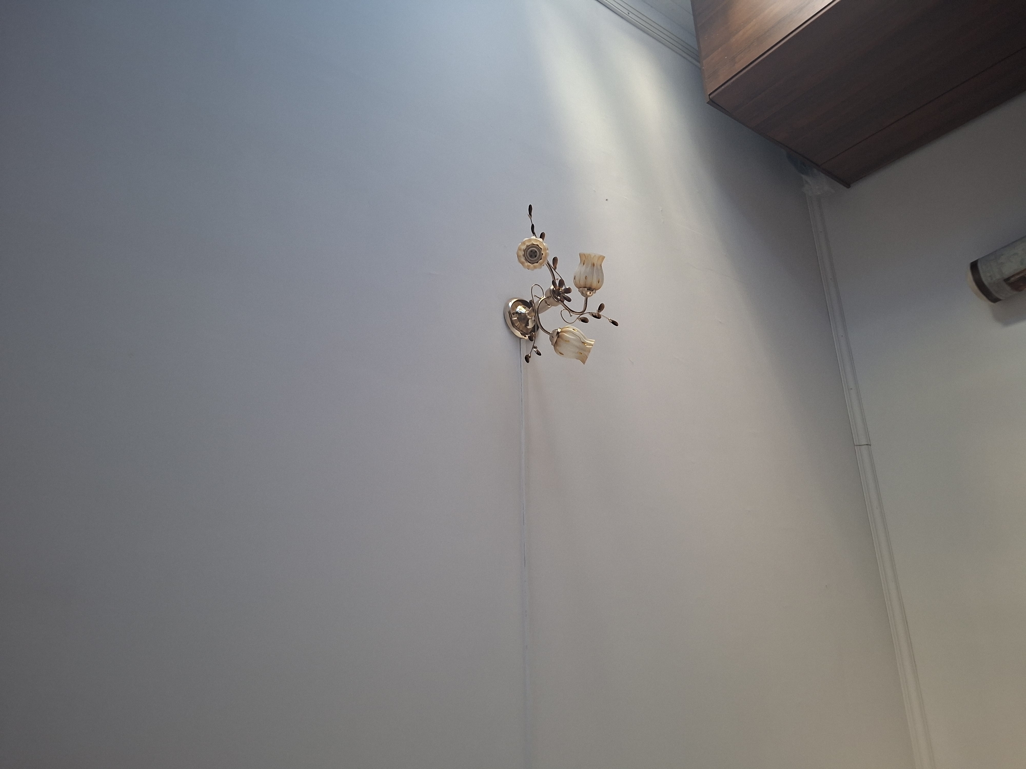 An indoor ceiling with a light fixture is shown. The light fixture is made of gold-colored metal and has three tulip-shaped glass shades. A dark brown cabinet is visible in the bottom right corner of the image, along with part of a rolled-up rug. The ceiling is white and there is a thin horizontal line across it. The location is the city of Zaşağan, Kazakhstan.