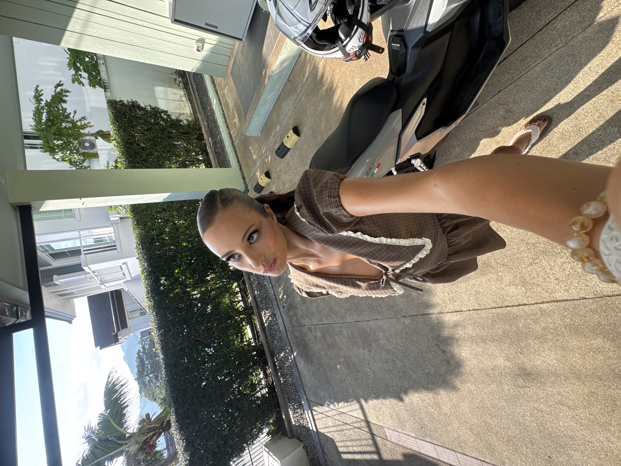 The image is a selfie taken by a woman in Chalong, Thailand, seemingly in the driveway of a residence. She is wearing a brown plaid dress with white lace trim and a beaded bracelet on her outstretched arm. A motorcycle with a helmet is parked beside her on the concrete driveway. In the background, there is a building with windows and green foliage. The day appears sunny, with shadows cast on the ground.