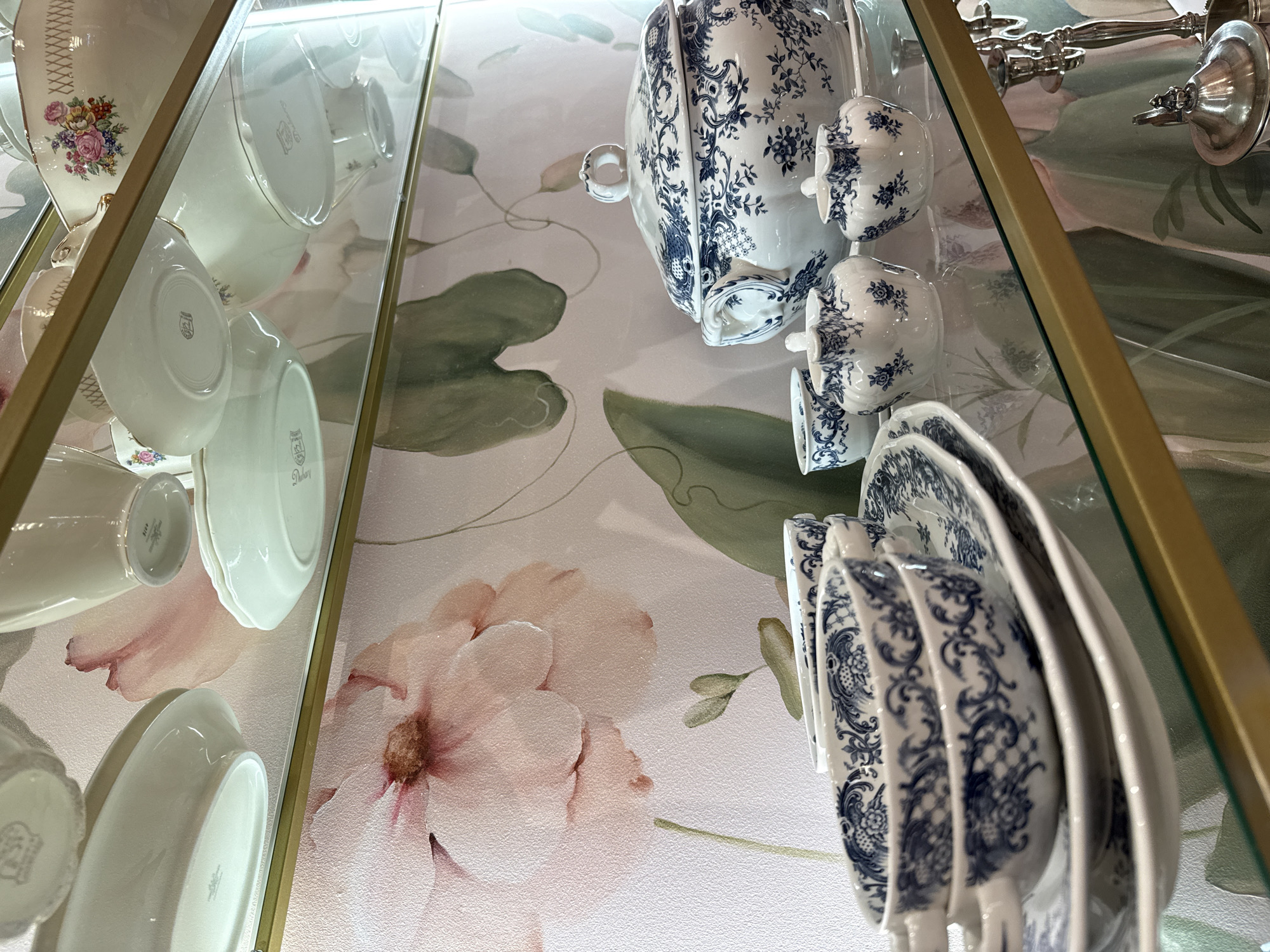 The image shows a close-up view of a display shelf with white porcelain dishes adorned with blue floral and scroll patterns. On the bottom shelf, two teacups are stacked, placed on a matching saucer. To the right of the teacups are two small creamers or sugar bowls, also with the blue pattern. Behind them is a large lidded tureen or serving bowl, decorated with the same blue motif. The background is a light-colored wallpaper featuring a large, soft-focus floral print with pink and green tones. The shelf itself is made of glass and edged with a gold-colored metal frame. The scene appears to be an indoor display, possibly in a home, shop, or museum. There are no people or animals visible. The lighting suggests an indoor environment with artificial light. There is no information to ascertain the location of this display in the city of Kyiv, Ukraine.