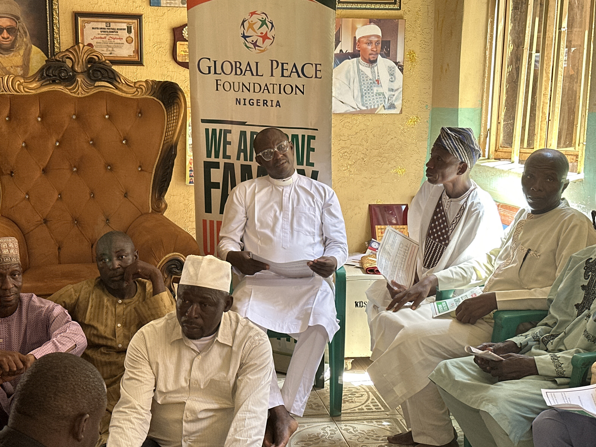 Here is a description of the image:
The image shows a gathering of men in what appears to be an office or meeting room, possibly in Unknown, Unknown, as indicated by the banner of the Global Peace Foundation Nigeria.
Several men are seated or squatting on the floor. One man, dressed in white, is seated on a green chair and holding papers in his hands, likely leading a discussion. Other men are wearing traditional or religious attire, including kufis and boubous.
In the background, there is a banner for the "Global Peace Foundation Nigeria," as well as framed pictures and certificates on the wall, including a "Football Diploma" from "NAFYA AMINA FOOTBALL ACADEMY SPORTS COMPLEX." The room's interior shows decorative wallpaper and patterned floor tiles. There's an ornate armchair on the left side of the scene.