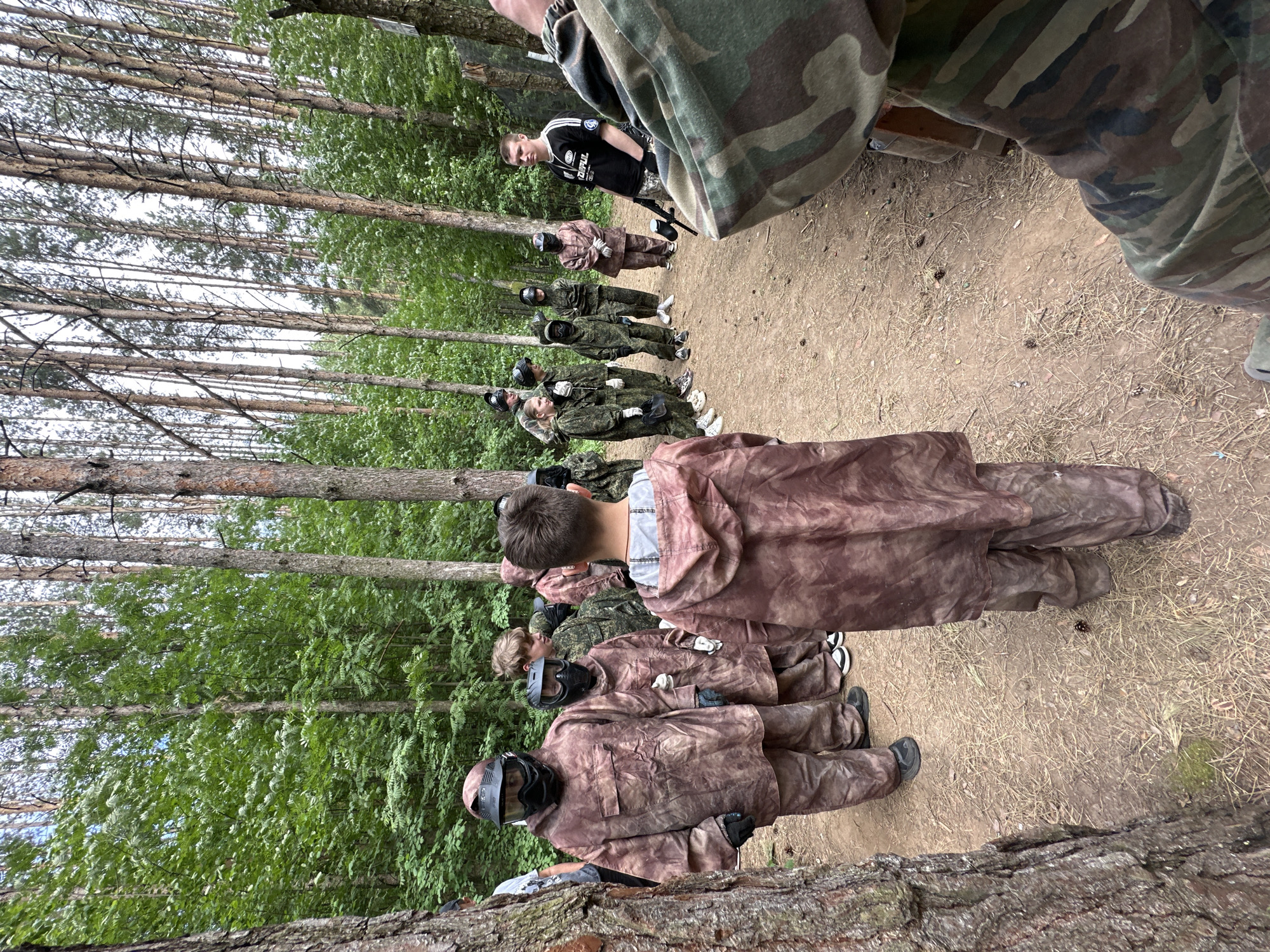 A group of approximately 10-12 people, mostly teenagers, are standing in a line along a dirt path in a wooded area. They are wearing camouflage clothing and protective face masks, suggesting they are participating in an activity like paintball or airsoft. The location appears to be a natural setting with tall trees lining the path. The presence of participants in camouflage suggests an outdoor recreational event or training. Without additional context, it's impossible to confirm the specific location within Votkinsk or provide further details about the activity or purpose.