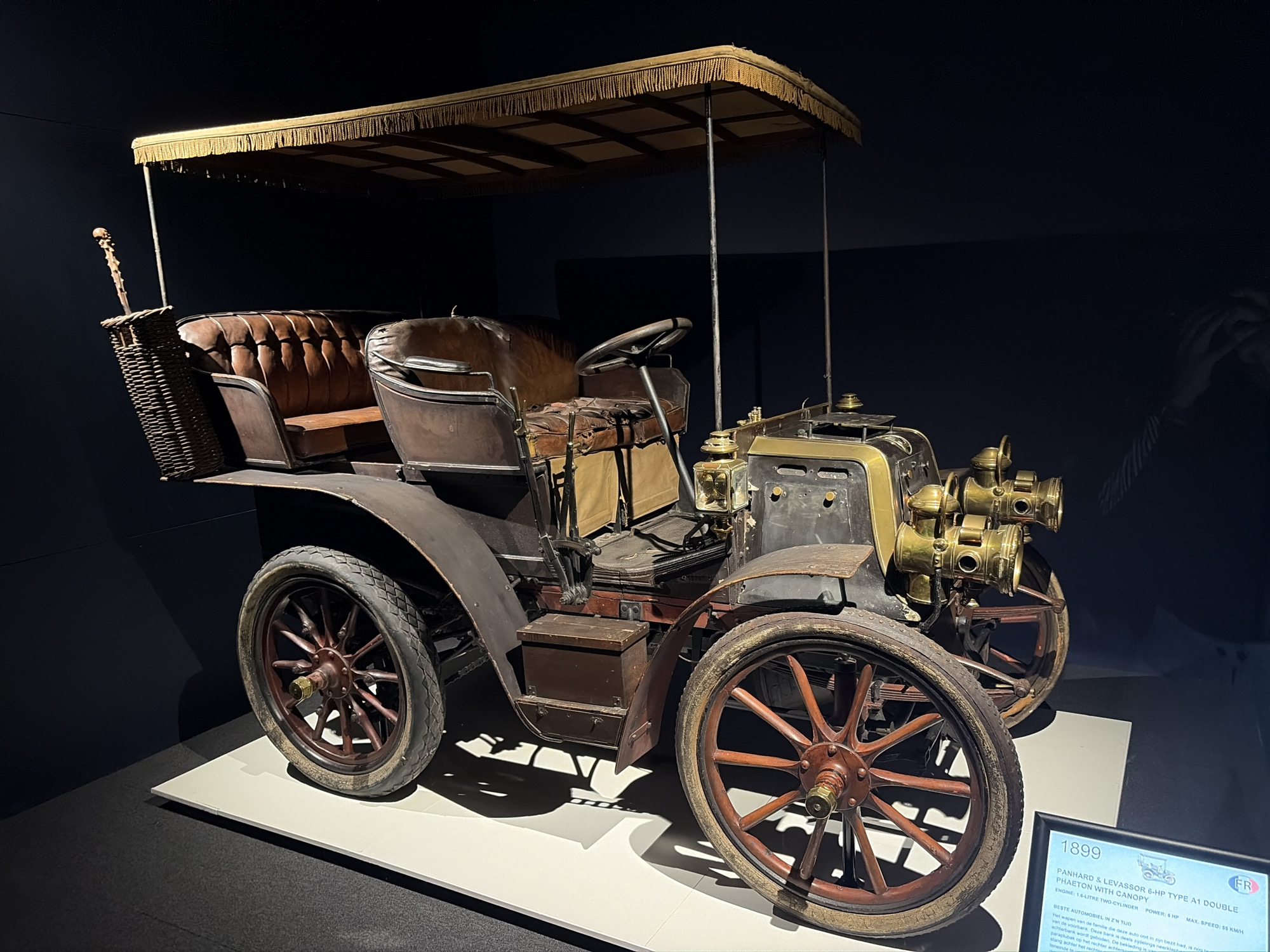 A vintage 1899 Panhard & Levassor 6-HP Type A1 Double Phaeton with Canopy car is prominently displayed indoors, likely in a museum or exhibition hall. The car, viewed from a three-quarter angle, rests on a white platform against a dark background, suggesting focused lighting on the exhibit.
The automobile features a light brown fringed canopy, supported by slender metal poles. Its body is dark, with rich brown tufted leather seats showing signs of age and wear. The driver's side features a simple black steering wheel. On the passenger side, a wicker basket is attached, holding a decorative stick with a skull-like or carved top. The front of the car is adorned with multiple brass headlights and other metallic fixtures, hinting at early automotive engineering. The wheels are made of dark wood with spokes and are fitted with rubber tires. A small wooden box is visible on the side step between the front and rear wheels.
An informational plaque next to the car provides details. Visible text reads: "1899 PANHARD & LEVASSOR 6-HP TYPE A1 DOUBLE PHAETON WITH CANOPY. ENGINE: 1.6-LITRE TWO-CYLINDER POWER: 6 HP MAX. SPEED: 55 KM/H." Below this are details in Dutch, including "BESTE AUTOMOBIEL IN Z'N TIJD" (Best automobile of its time) and descriptions of the car's features like a foldable front seat and an umbrella stand at the rear. The overall setting is calm and focused on presenting the historical vehicle.
