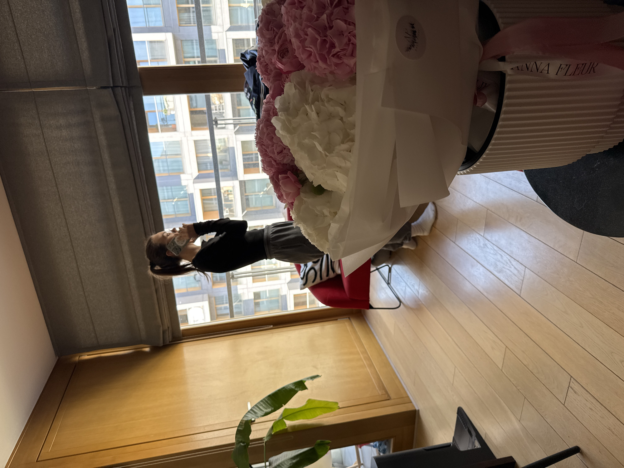 A woman wearing a mask stands indoors, partially framed by a window in what seems to be an apartment or office building, possibly located in Warsaw, Poland, as suggested by the architectural style of the building visible through the window. The woman, dressed in casual clothing, is holding a mobile phone and appears to be taking a photo of a large bouquet of pink and white flowers, which are the focal point. The flowers are presented in a tall, white, corrugated container with a branded ribbon indicating "ANNA FLEUR." The room has wooden flooring, a red chair, and partial views of a plant and television. The window offers a view of a modern building with many windows. A grey curtain or blind is visible to the left of the window.