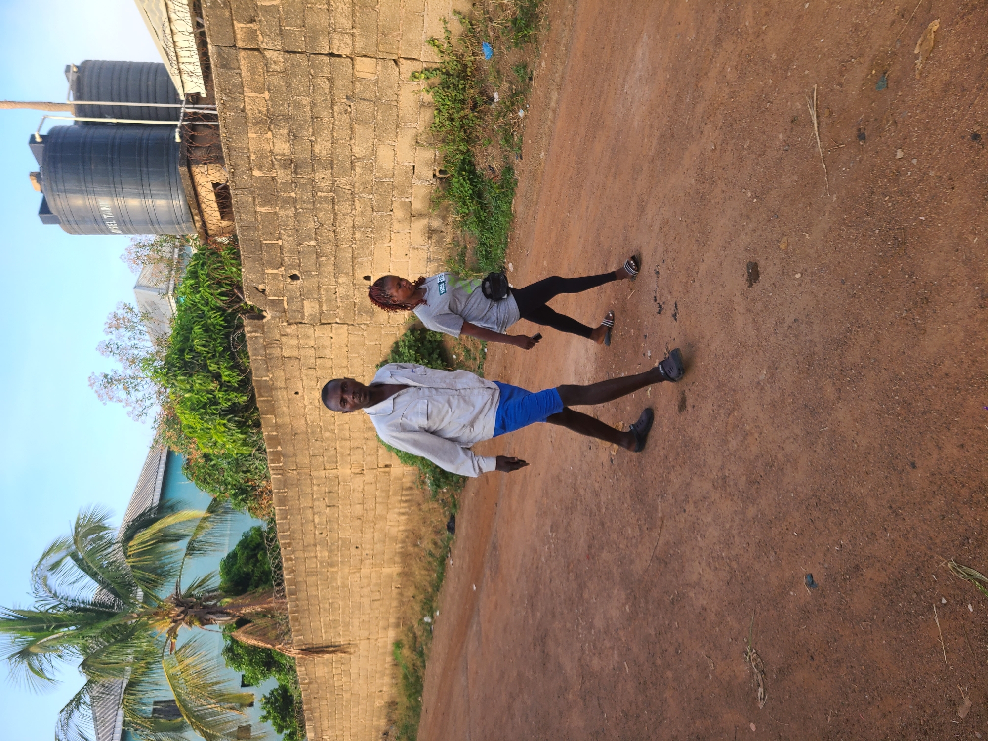 Two people stand on a dirt road next to a brick wall. The person in the foreground wears a light-colored shirt and blue shorts. The second person wears a light-colored shirt and dark pants. Behind the wall are trees and buildings. Water tanks are visible atop the building. The location is in Jos, Nigeria, based on the presence of distinctive tropical vegetation.