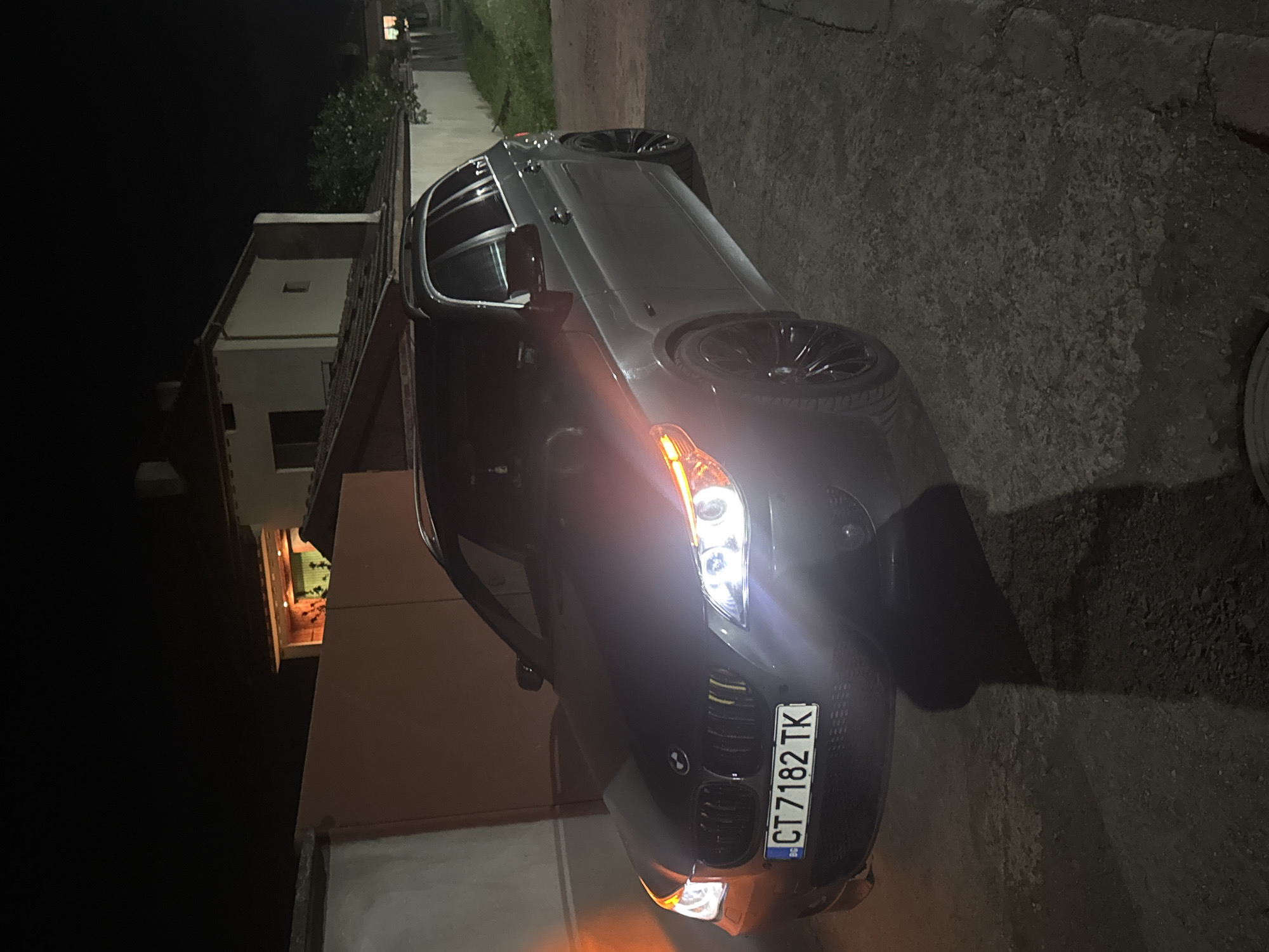 A gray BMW sedan is parked on an asphalt surface in an outdoor environment at night. The car's headlights are illuminated. A house is visible to the left of the car, and vegetation is seen in the background. The license plate reads "CT 7182 TK". The location is Sarnevo, Bulgaria based on the license plate.