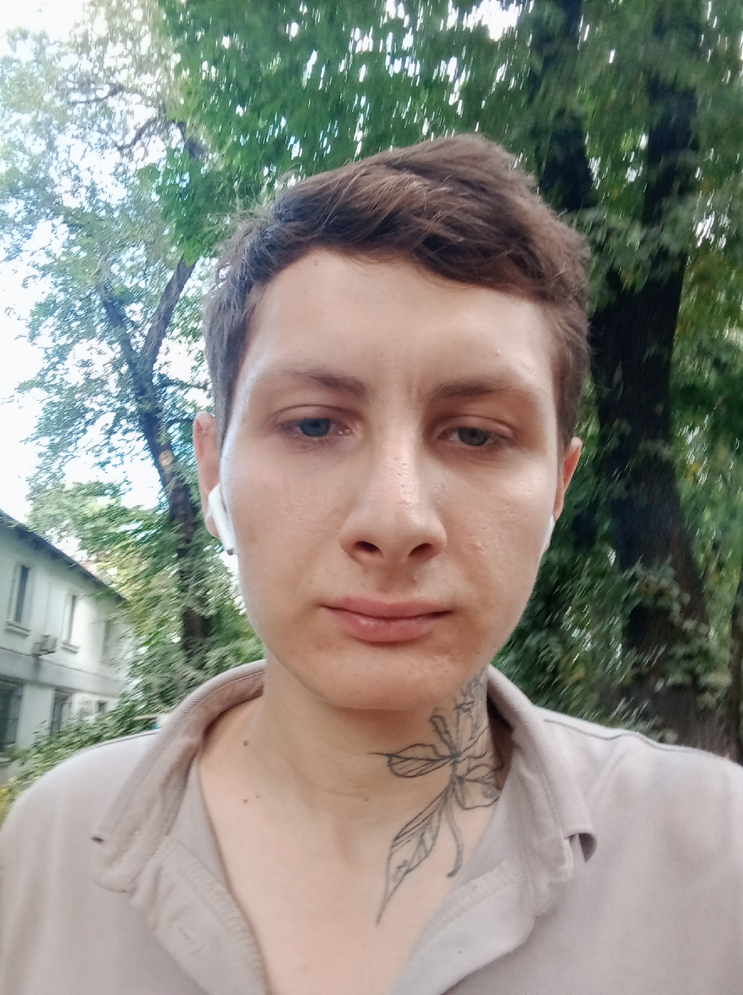 A close-up, waist-up shot shows a person with short brown hair, light skin, and blue eyes. They are wearing white earbuds and a beige polo shirt. A tattoo of a flower with leaves is visible on the left side of their neck. The person is looking directly at the camera. In the background, there are trees with green leaves and a white building with windows, suggesting an outdoor urban setting. The scene is consistent with a residential area in Almaty, Kazakhstan, possibly near a park or a courtyard of an apartment building. The lighting suggests it is daytime.