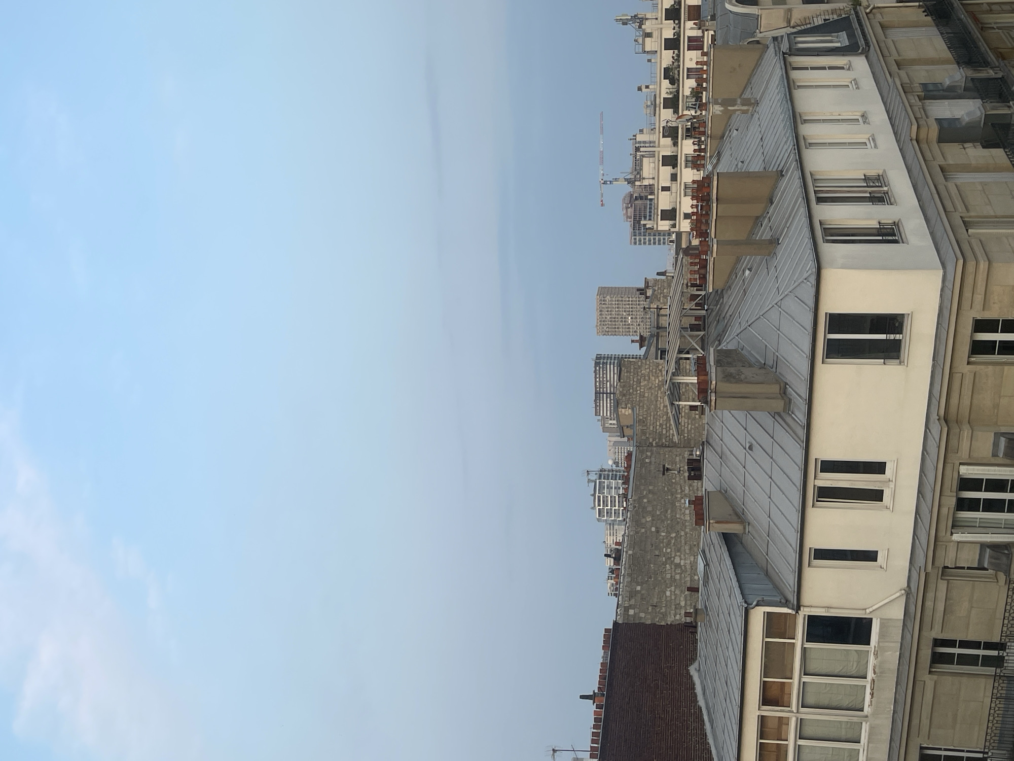 This is an image of a cityscape in Paris, France, taken from a high vantage point. The sky is a pale blue with some wispy clouds, suggesting it might be early morning or late afternoon. In the foreground, there are several Parisian-style buildings with mansard roofs, characterized by their grey metal sloping surfaces and numerous chimneys. These buildings feature cream-colored facades and numerous windows, some of which are large and multi-paned, while others are narrower and taller.
In the middle ground and background, more buildings are visible, including some taller, modern structures and older stone buildings. Construction cranes are also visible in the distance, indicating ongoing development in the city. The overall impression is one of a densely populated urban environment. There are no people or animals visible in the frame.