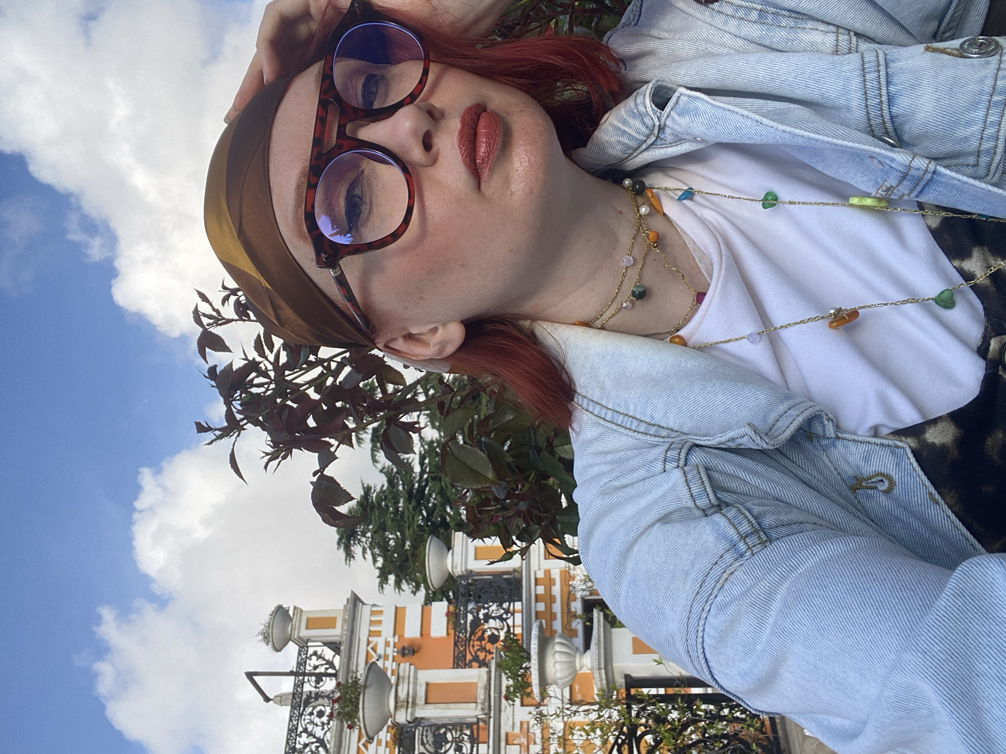 A woman with red hair, wearing glasses and a headscarf, poses outdoors. She is wearing a white t-shirt and a light blue denim jacket, accessorized with two beaded necklaces.
The background reveals architectural details, including a building with an orange and white facade. The presence of ornate ironwork and plantings suggests a refined outdoor setting. In the distance, a blue sky with fluffy white clouds is visible. The overall setting, based on the architectural style and general appearance, suggests that the location is the city of Sochi, Russia.