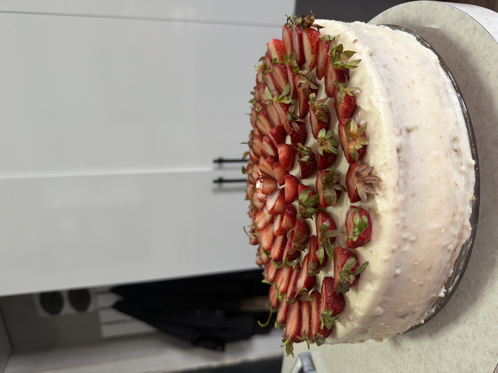 A close-up, angled shot depicts a white-frosted cake decorated with sliced strawberries, situated indoors on a countertop or similar surface. The cake is positioned on a cake stand. In the background to the left is a white cabinet. A dark object, possibly an umbrella, is partially visible under the cabinet. The image does not provide enough information to determine the exact location in Tbilisi, Georgia. There are no people visible in the image. The primary subject is the cake and its strawberry decoration.