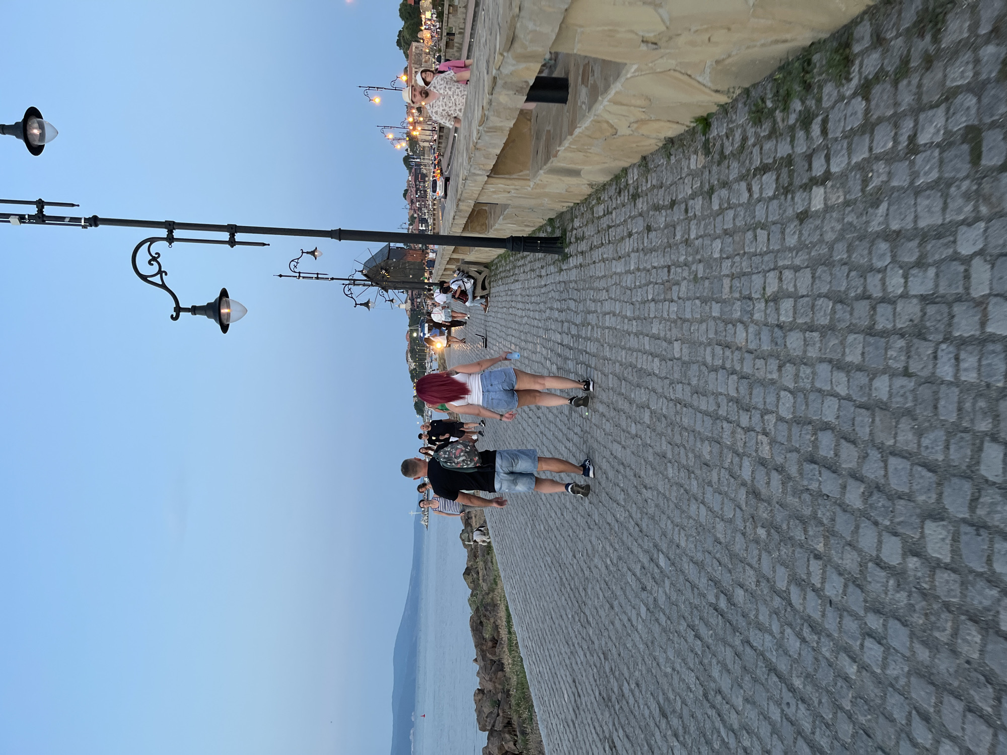 In Nesebar, Bulgaria, a paved promenade runs alongside the sea. In the foreground, a man and a woman walk away from the camera on the cobblestone path. The man wears a black t-shirt and denim shorts, carrying a patterned backpack. The woman has red hair and wears a white tank top with denim shorts. Numerous streetlights, some lit, line the promenade. Further down, other people are visible strolling, sitting on benches, and interacting. Buildings, including what appears to be a windmill, and a line of streetlights, are visible in the distance along the coastline. The sea stretches to the left, with a distant coastline on the horizon. The sky is a clear, pale blue, suggesting it is either early morning or late evening. A stone wall with arched openings borders the right side of the promenade.