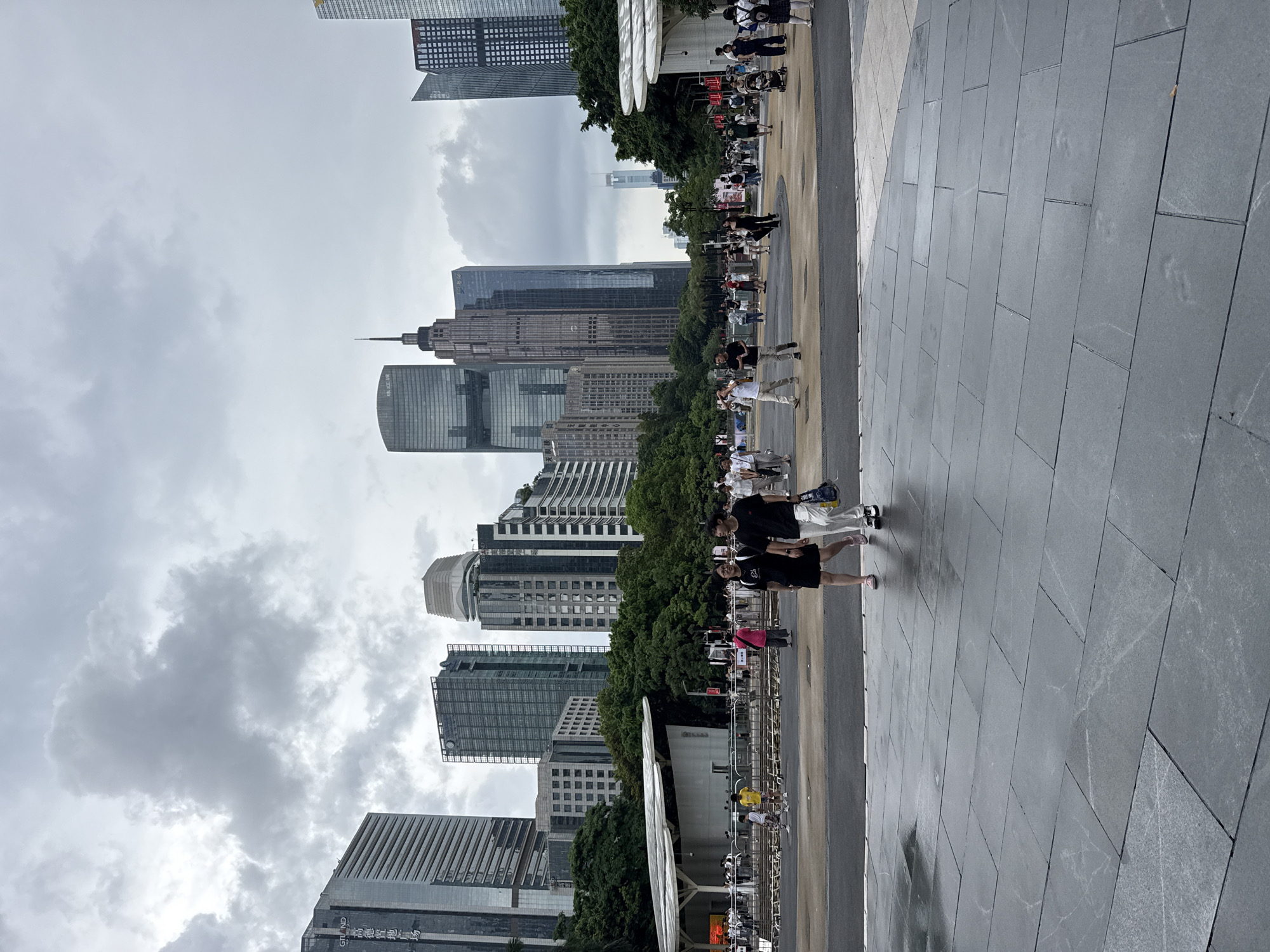 This media file depicts a street scene in Tianhe District, China, on an overcast day. The foreground is dominated by a wide, gray tiled walkway. Several people are walking or standing on this walkway, some carrying bags. Behind the walkway, a line of lush green trees forms a verdant border. Beyond the trees, a collection of modern skyscrapers rises into the cloudy sky, showcasing a dense urban landscape. The buildings are of varying heights and architectural styles, some featuring glass facades. A few individuals are visible near the trees and in the distance, suggesting a public space with pedestrian activity. The overall atmosphere appears calm and uneventful, with no overt indications of a specific event or strong emotions. The cloudy weather suggests it could be morning or afternoon.