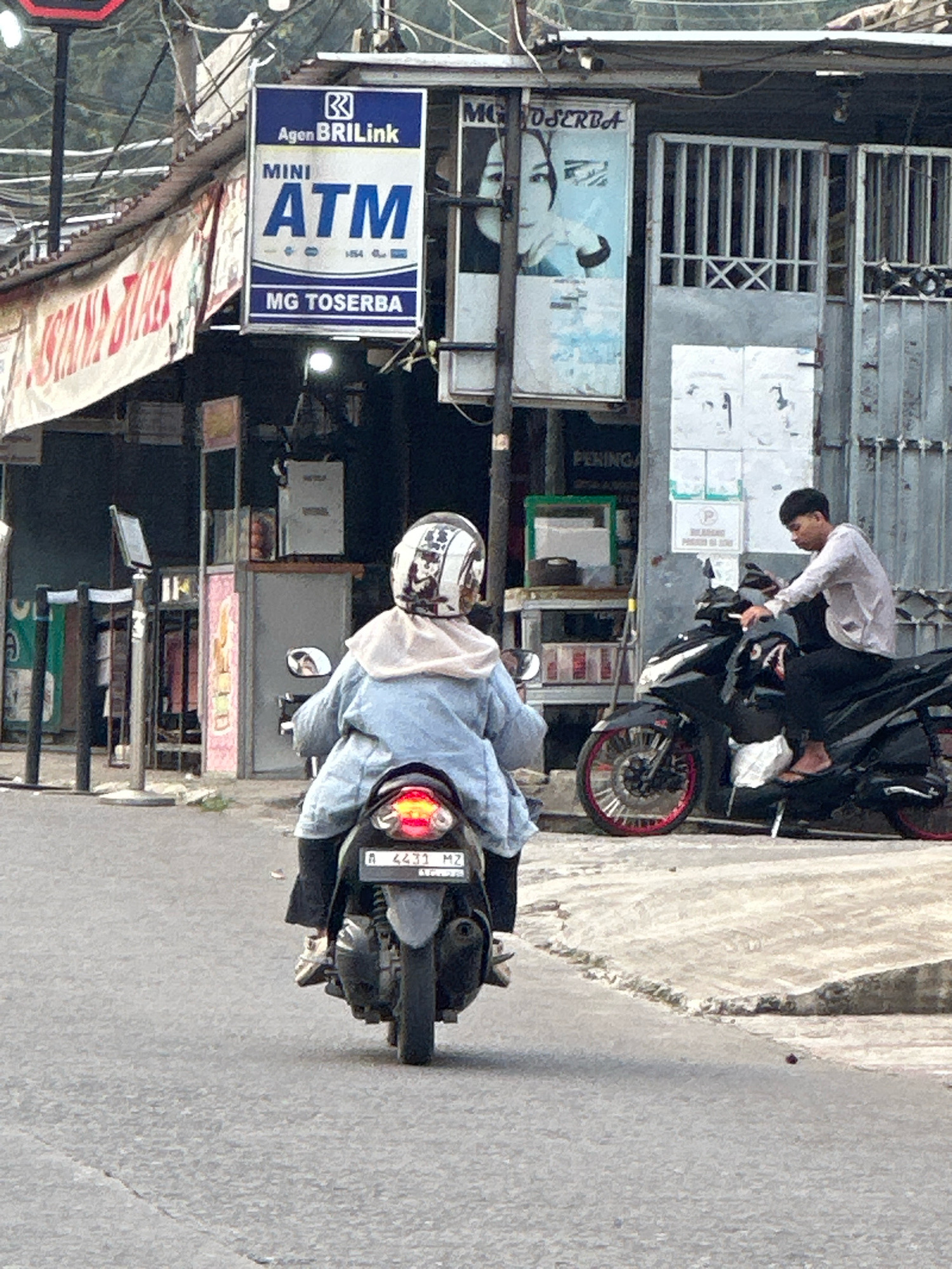 The image depicts a street scene in Saruni, Indonesia. In the foreground, a person wearing a light blue denim jacket and a white helmet is riding a scooter. The scooter has a license plate that reads "A 4431 MZ". Behind the rider, a black motorcycle is parked with a person wearing a light-colored shirt and dark pants sitting on it. To the left of the frame, a sign for "Agen BRIlink MINI ATM" is visible. Further back, a building with metal gates is present. The environment appears to be an urban street with some shops and signage.