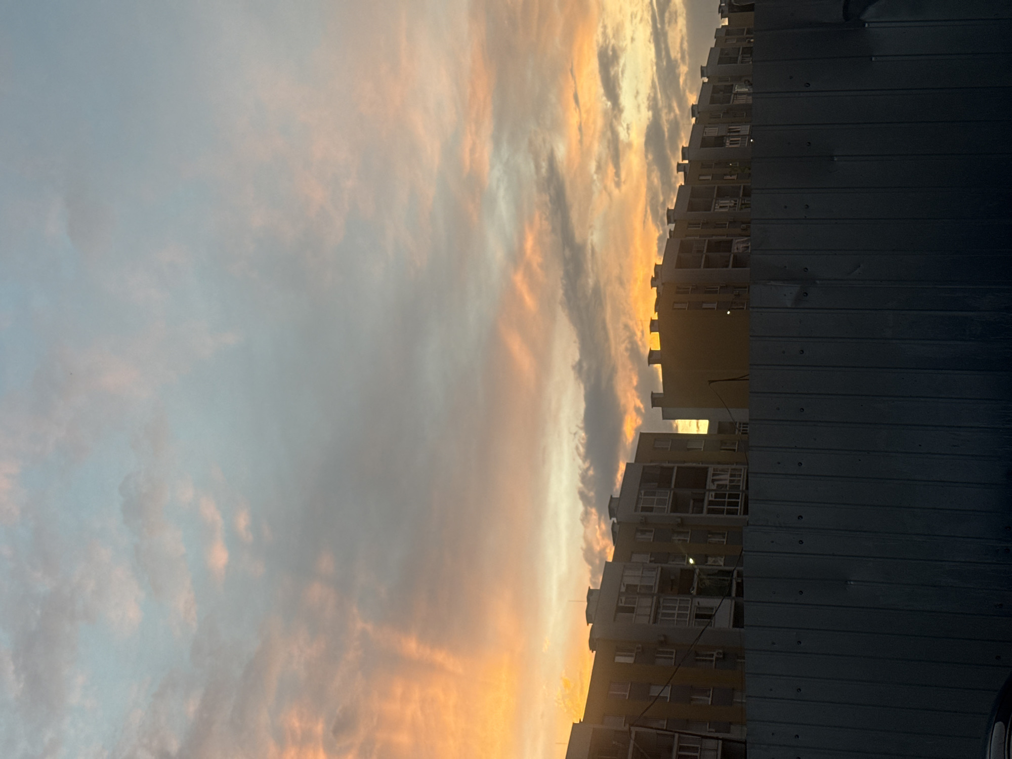 The image depicts a sunset over a residential area in Avanhard, Ukraine. The sky is filled with clouds displaying hues of orange, pink, and blue, indicative of the twilight hour. In the foreground, a dark, corrugated metal fence runs horizontally across the bottom half of the frame. Behind the fence, several multi-story apartment buildings are visible, their facades illuminated by the fading sunlight. The architecture of the buildings suggests a post-Soviet style, with repetitive window patterns and simple, functional designs. The scene is devoid of any people or discernible activity, presenting a still, urban landscape at the close of day. The lighting is soft and diffused, characteristic of a sunset.
