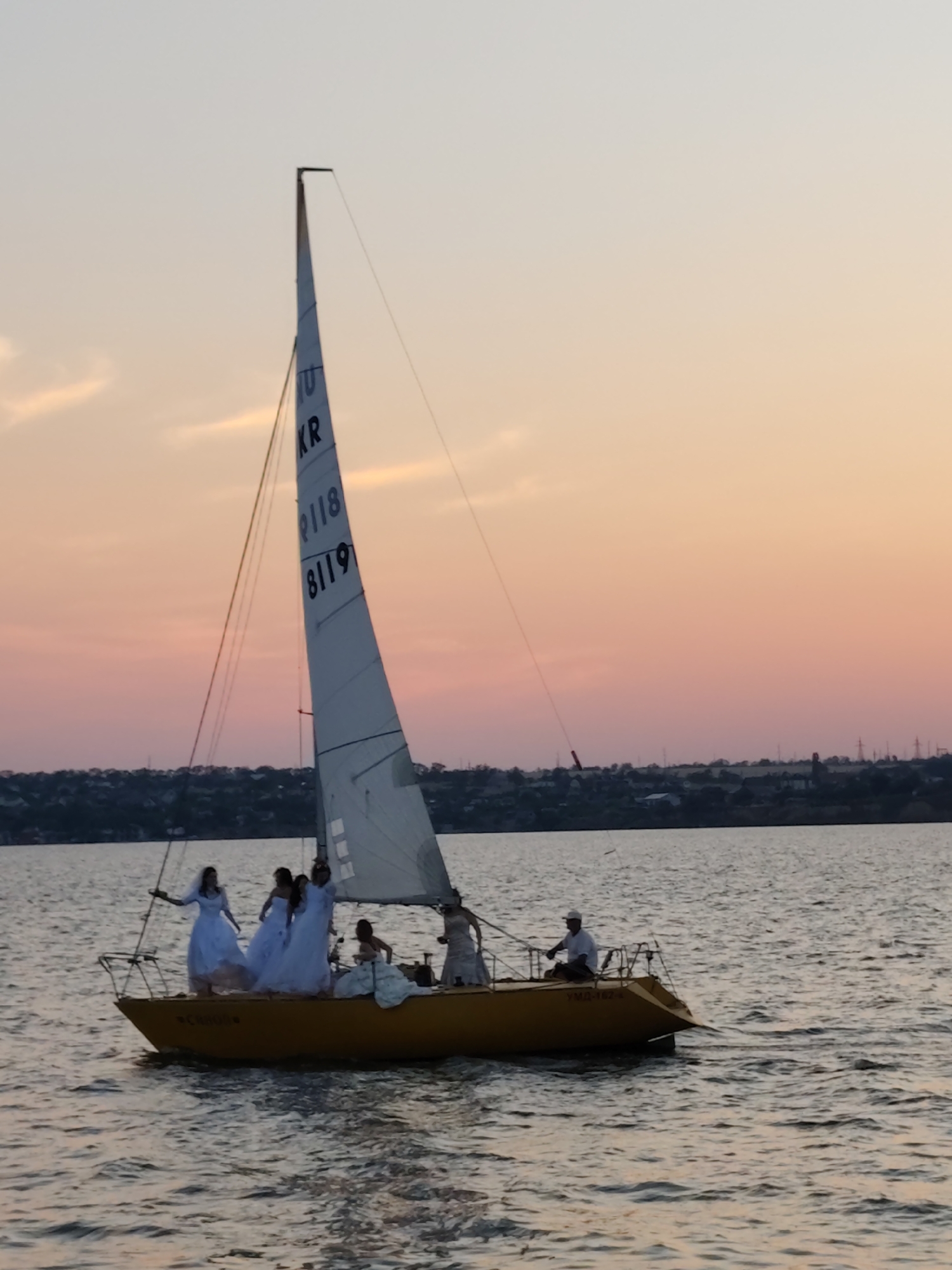 This media file depicts a group of people on a sailboat during sunset. The sailboat is yellow with a white sail that has black numbers and letters on it. There are at least six people on the boat, most of whom appear to be women wearing white dresses. One person, presumably the captain, is steering the boat. The sky is a gradient of orange, pink, and light blue, indicating it is either sunrise or sunset. The water is calm, with gentle ripples. In the background, there is a distant shoreline with trees and buildings, and some utility poles are visible. The overall mood is serene and perhaps celebratory. The text visible on the sail reads "10", "KR", "8118", and "8119". There is also faint text on the side of the boat, partially obscured by shadows and the angle. Based on the location context provided, this scene is likely taking place in Mykolaiv, Ukraine.