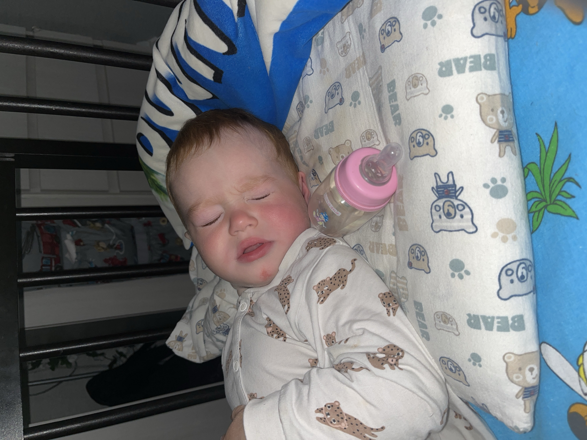 A baby is sleeping in a crib. The baby is wearing a white pajama with a leopard print. There is a pink baby bottle next to the baby. The crib has black bars. The bedding has a bear print and a blue and white striped pattern. The image appears to be taken indoors. No specific location in Chaikovsky, Russia can be determined from the image.