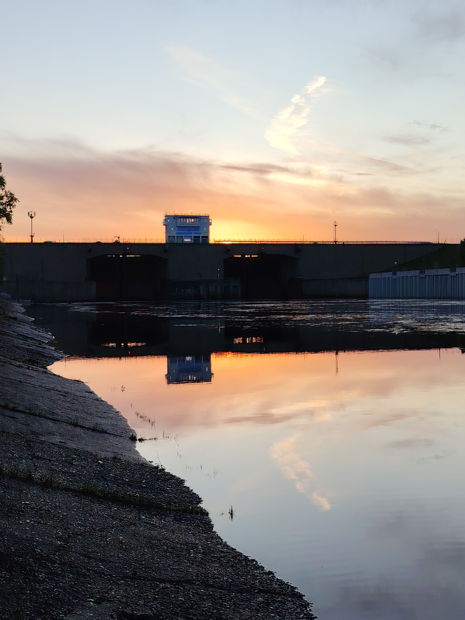 The image shows a dam in Chaikovsky, Russia at sunset. The sky is a mix of orange, pink, and blue. The dam is silhouetted against the sky, and its reflection is visible in the water. The foreground is a rocky shoreline. The sky shows a trail. No people are visible.