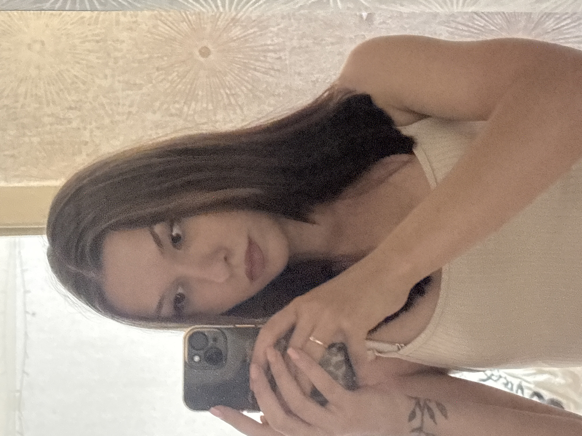 A young woman with dark brown hair is taking a mirror selfie indoors. She is wearing a light-colored tank top. Her left arm is holding a smartphone, and a ring is visible on her left ring finger. A tattoo of leaves is visible on her left forearm. The background appears to be a wall with patterned wallpaper, possibly in a residential setting. There is no indication of the specific location within the city of Inskoy, Russia.