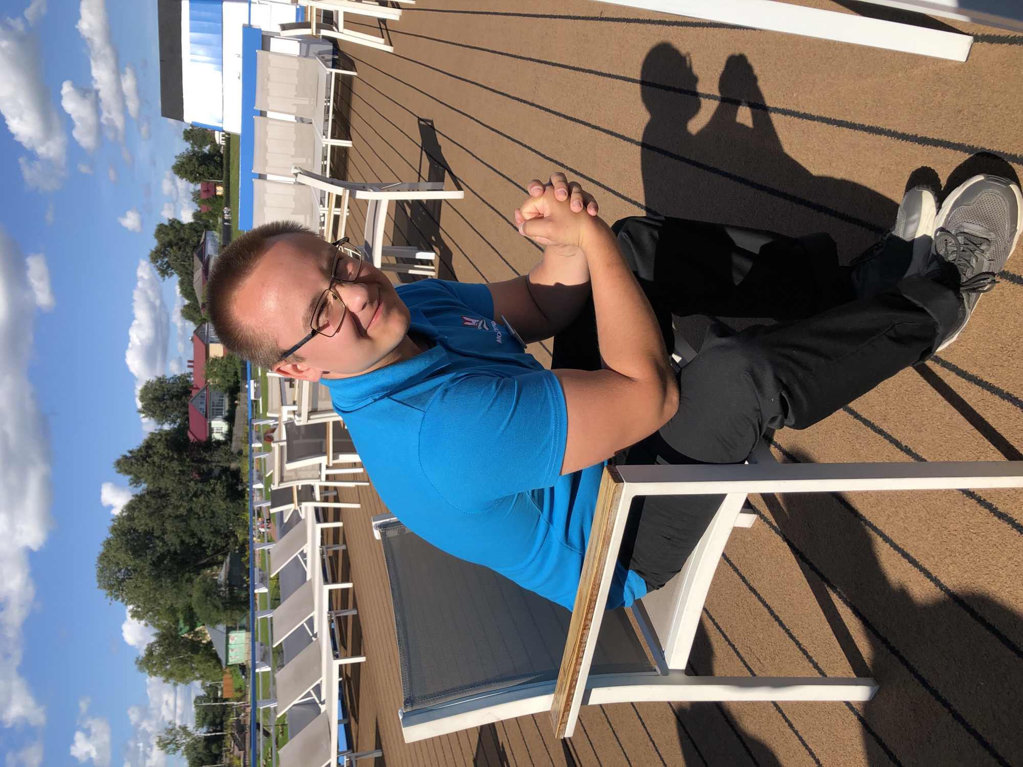 A young man with short brown hair, glasses, and a blue polo shirt is sitting in a white outdoor chair on a wooden deck. He is looking at the camera with a slight smile and has his hands clasped in his lap. In the background, there are rows of lounge chairs and buildings that appear to be in a rural or suburban setting. The sky is blue with white clouds. The location is identified as Myshkin, Russia.