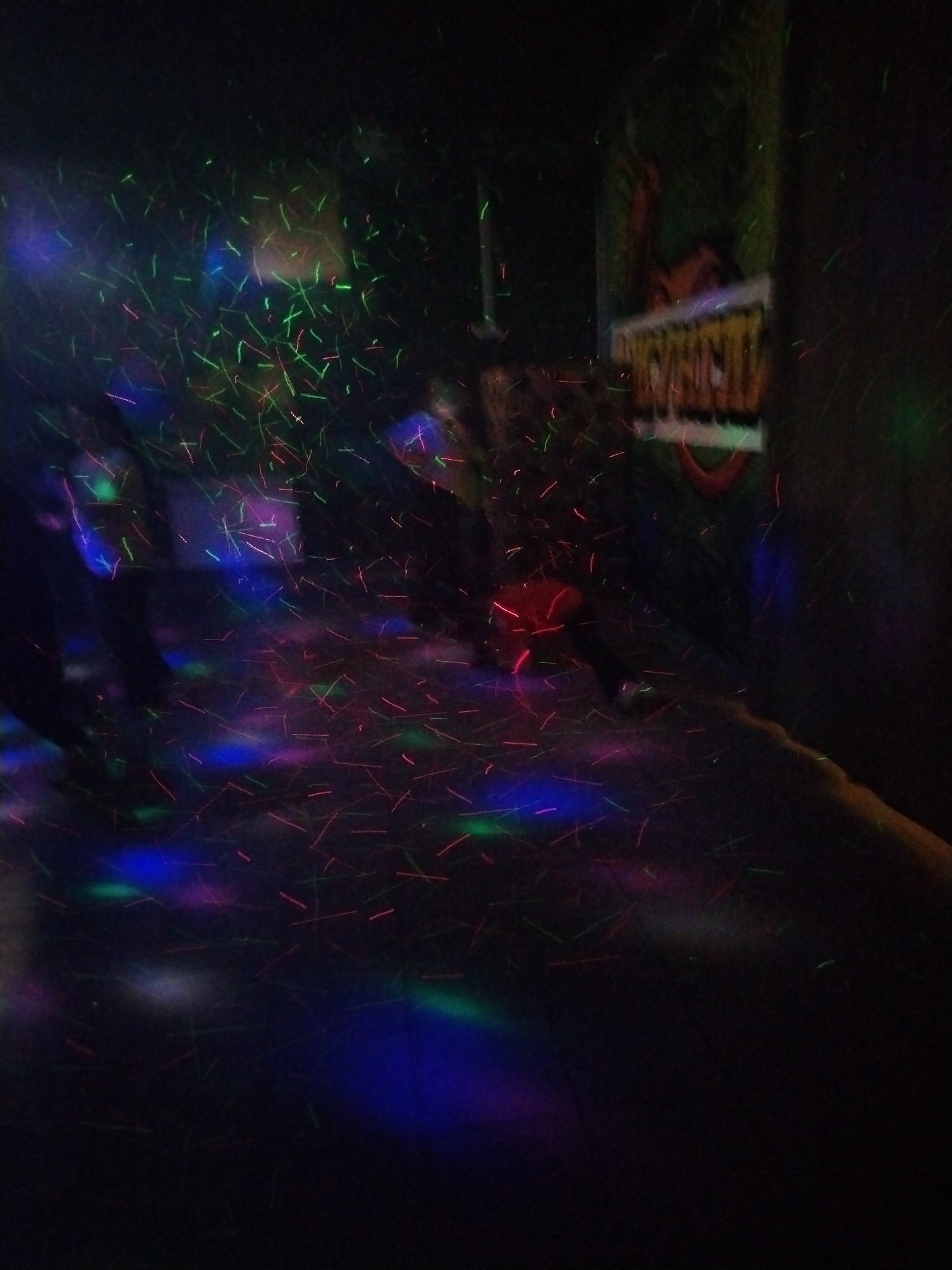 The image depicts a dimly lit interior, possibly a laser tag arena or entertainment center, located in Rusanovo, Russia. Multiple people are partially visible in the midground, their forms obscured by the low light and laser effects. A sign is partially visible on the right. The environment is filled with streaks of red, green, and blue laser lights, creating a dynamic and chaotic visual effect. The floor also reflects these laser lights. The overall impression is one of activity and entertainment in a dark, enclosed space.