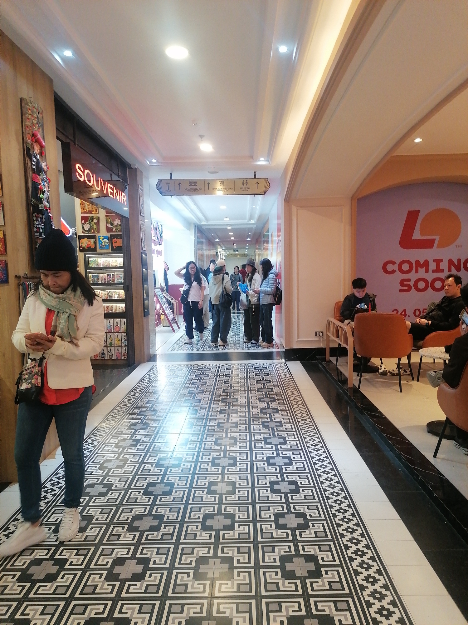 The image shows an indoor scene in Sa Pa Ward, Vietnam. A woman wearing a black beanie, white jacket, red shirt, and jeans stands to the left, looking at her phone. She has a floral handbag on her shoulder. Behind her is a souvenir shop. In the center, people are walking down a hallway. To the right, there's a seating area with people sitting in orange chairs. A sign on the wall reads "LO COMING SOON 24.05". The floor is tiled in a geometric pattern. The overall environment suggests a commercial or shopping center interior.