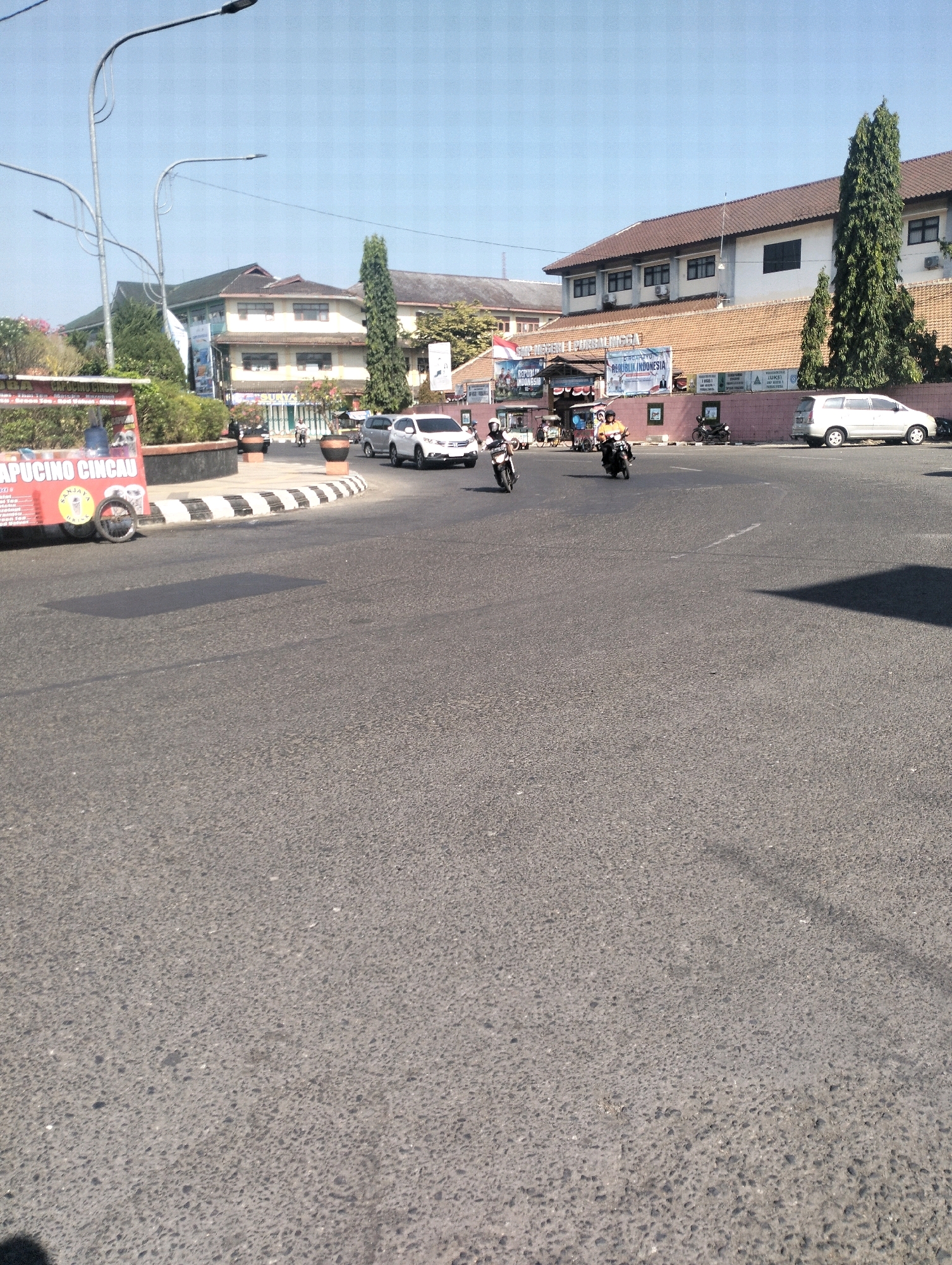 A street scene in Purbalingga, Indonesia, shows traffic on a sunny day. Two motorcycles are visible on the road, one carrying a rider wearing an orange shirt and helmet, and another with a rider in a black shirt and helmet. In the background, a white car is parked on the side of the road. Buildings line the street, including one with a brown tiled roof and multiple windows, and another with a light-colored facade and darker accents. A street cart with red and white signage is also present on the left side of the image. The sky is clear and blue.