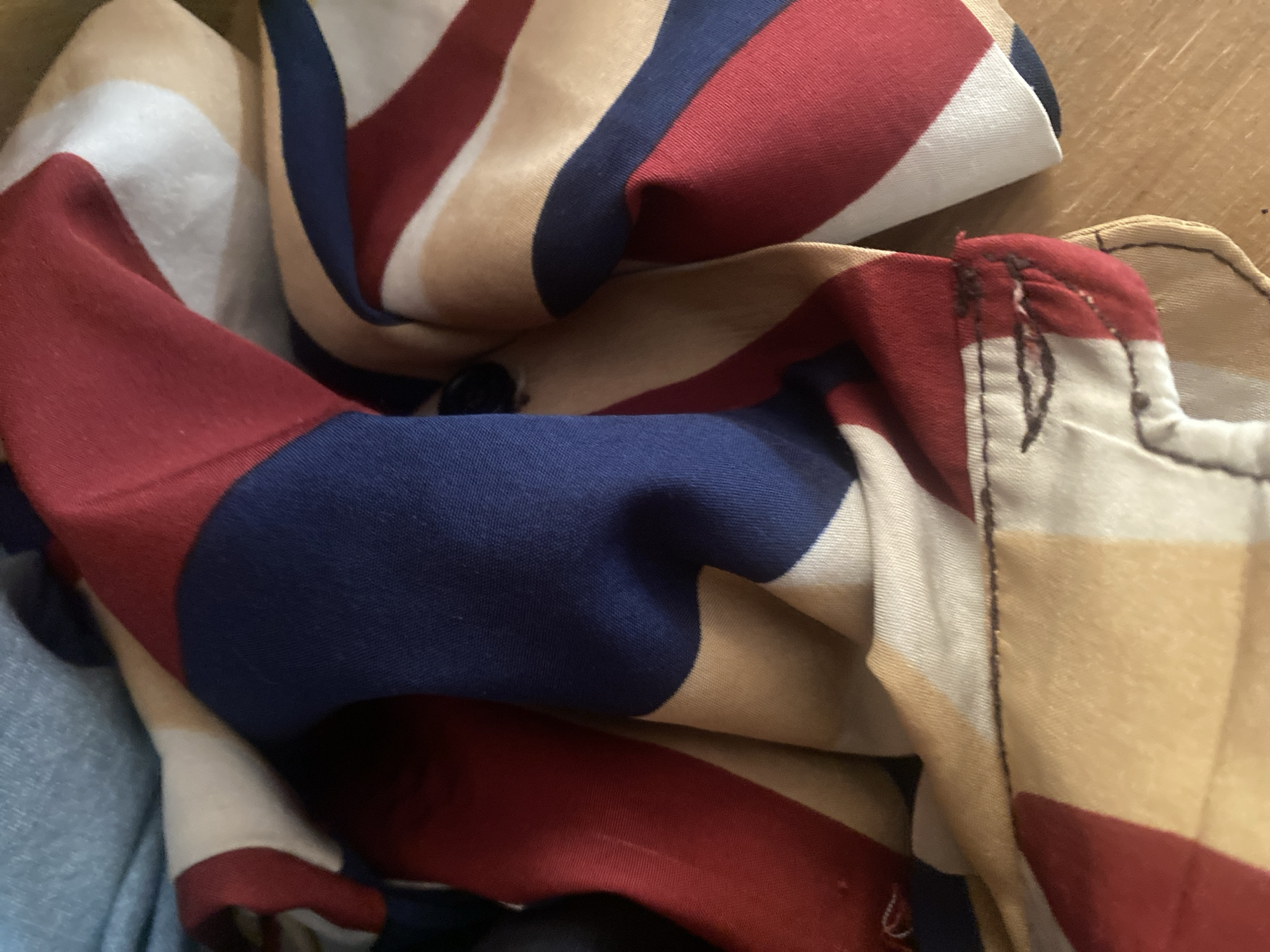 The image shows a close-up, cluttered view of a pile of fabric. The fabric has a striped pattern featuring bold, diagonal bands of red, navy blue, tan, and white. The material appears to be soft, possibly a light fabric like cotton or polyester, and is folded and crumpled, obscuring much of its entirety.
In the foreground, a section of navy blue fabric is visible, with a prominent wave-like fold. To the left, a strip of red fabric is draped over a lighter colored section. Further back and to the right, the fabric is layered, with the tan and white stripes predominantly showing, alongside more red and navy. There are hints of stitching and unfinished edges visible on some of the fabric pieces, suggesting they might be part of a garment, home textile, or craft project.
The lighting is somewhat dim, casting soft shadows and highlighting the texture of the fabric. The overall impression is one of disarray or being in storage. There is no clear indication of a specific activity or event taking place, nor are there any visible text, people, or discernible location cues within Jalingo, Nigeria. The focus is entirely on the patterned fabric itself.