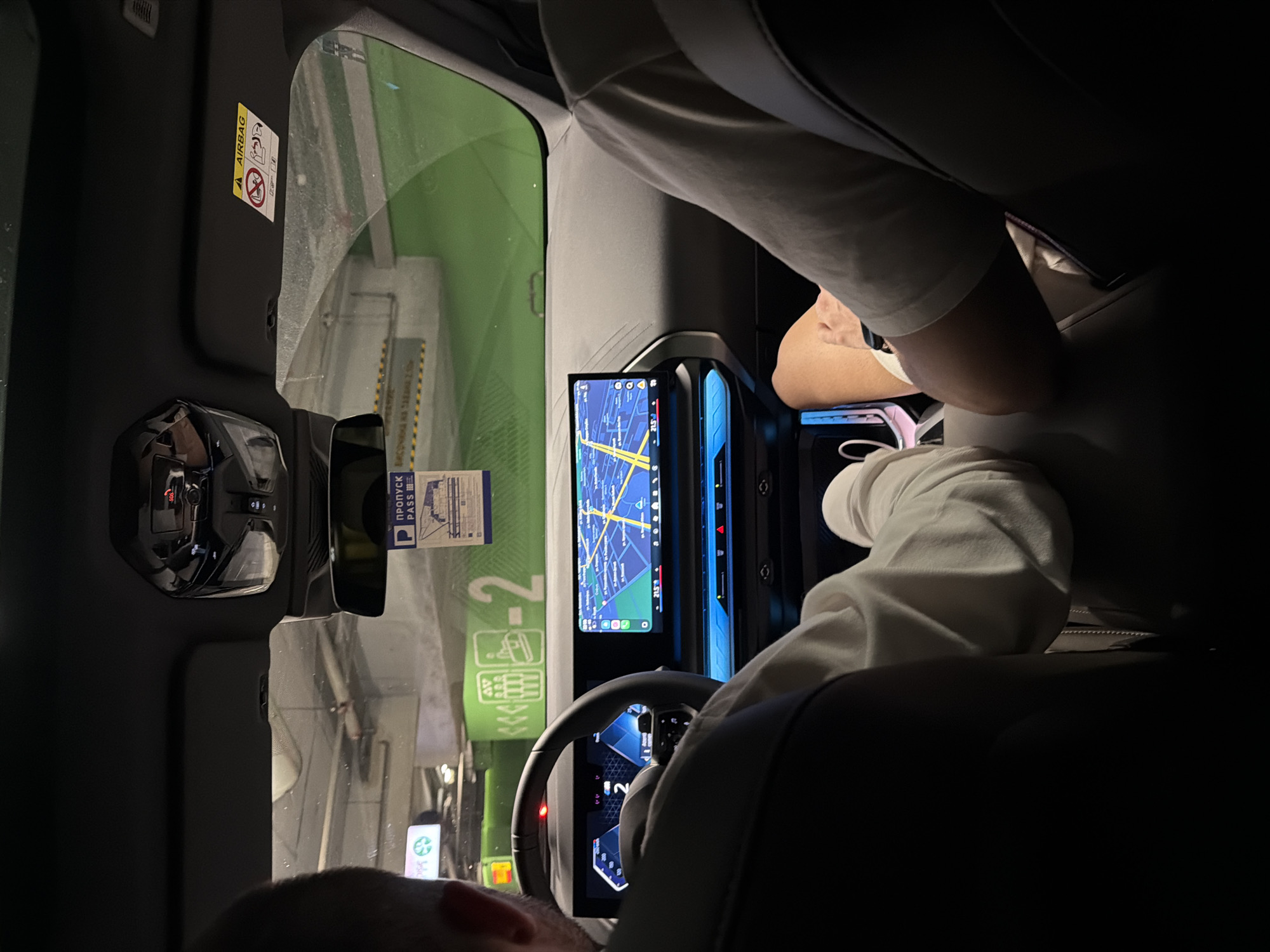 The image depicts the interior of a modern vehicle, specifically the front cabin, viewed from a rear passenger seat perspective. The vehicle is situated within a multi-story or underground parking garage in Sofia, Bulgaria.
Two adult males occupy the front seats. The driver, positioned on the left, is seen from behind the head and shoulder, wearing a light-colored long-sleeved top. His hands are placed on the steering wheel, behind which a digital instrument cluster displays a "2" prominently. The front passenger, on the right, is visible from the right arm and leg; he wears a light-colored short-sleeved top and dark shorts, with a smartwatch on his right wrist.
The dashboard features a continuous curved digital display unit. The central section of this display shows a navigation map with a city layout, indicating a temperature of "21.5°C." Blue ambient lighting is visible along the lower portion of the dashboard and center console. An overhead console contains functional buttons, including one marked "SOS." The passenger-side sun visor displays an "AIRBAG" warning sticker. A rectangular parking pass, featuring a blue "P" symbol and the Cyrillic word "ПРОПУСК" (PASS), hangs from the rearview mirror.
Through the windshield, the concrete structure of the parking garage is visible. Prominent green wall sections display a large white number "2" and various white graphical symbols, likely indicating parking levels or zones. A partially visible sign with a green recycling symbol is present in the upper left of the exterior view. The scene suggests the vehicle is stationary within the parking facility.