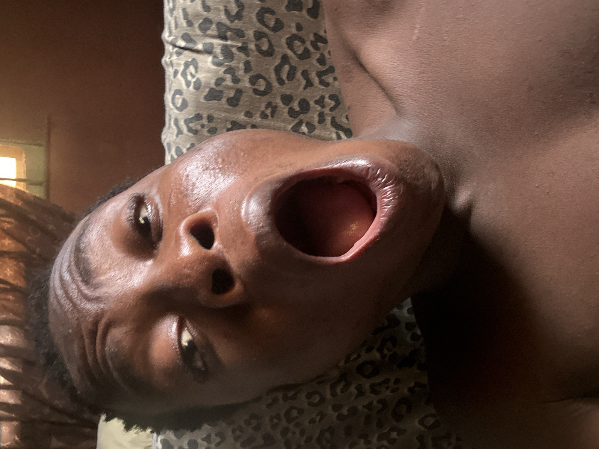 A close-up, low-angle shot depicts a person of color lying on a leopard-print surface. The person's face is contorted with their mouth wide open. The background shows a window with light coming through. No city or location is discernible from this image.