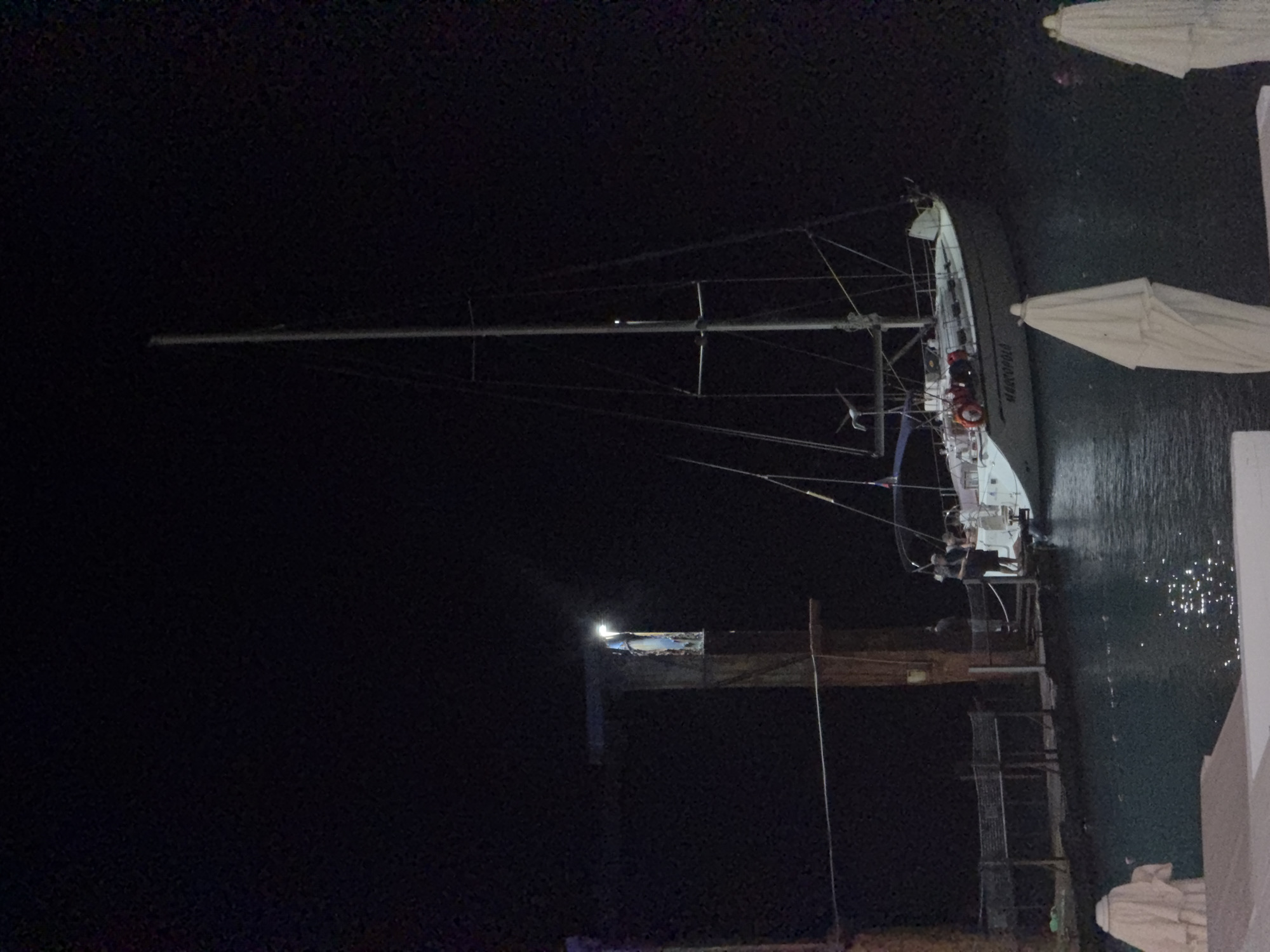 At night in Sochi, Russia, a white sailboat named "MARCOPOLO" is docked near a pier. Several individuals are visible on the deck of the sailboat and on the pier. A tall mast with rigging is prominent on the sailboat. The water reflects the light from the scene. In the foreground, parts of lounge chairs and white umbrellas are visible, suggesting a resort or recreational area. A lamppost illuminates a section of the pier.