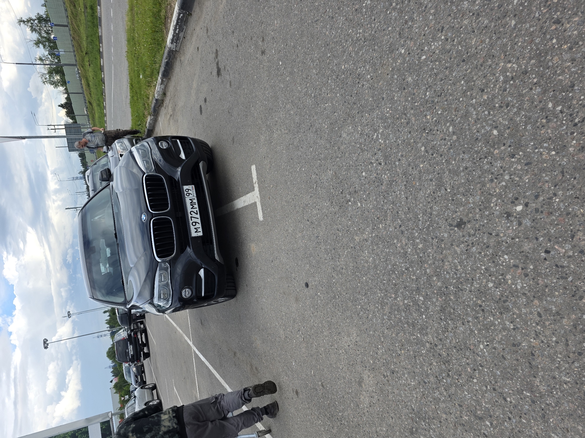 In this image, a man is seen leaning over the hood of a black BMW parked in a lot. He is wearing gray pants and a gray shirt. The car has a license plate that reads "M972MM99". Behind the BMW, several other cars are parked in a line, also facing the same direction. The scene appears to be an outdoor parking lot on a cloudy day, with streetlights visible in the background. The ground is asphalt, marked with white parking lines. The image is oriented vertically, with the sky and trees visible on the left side.